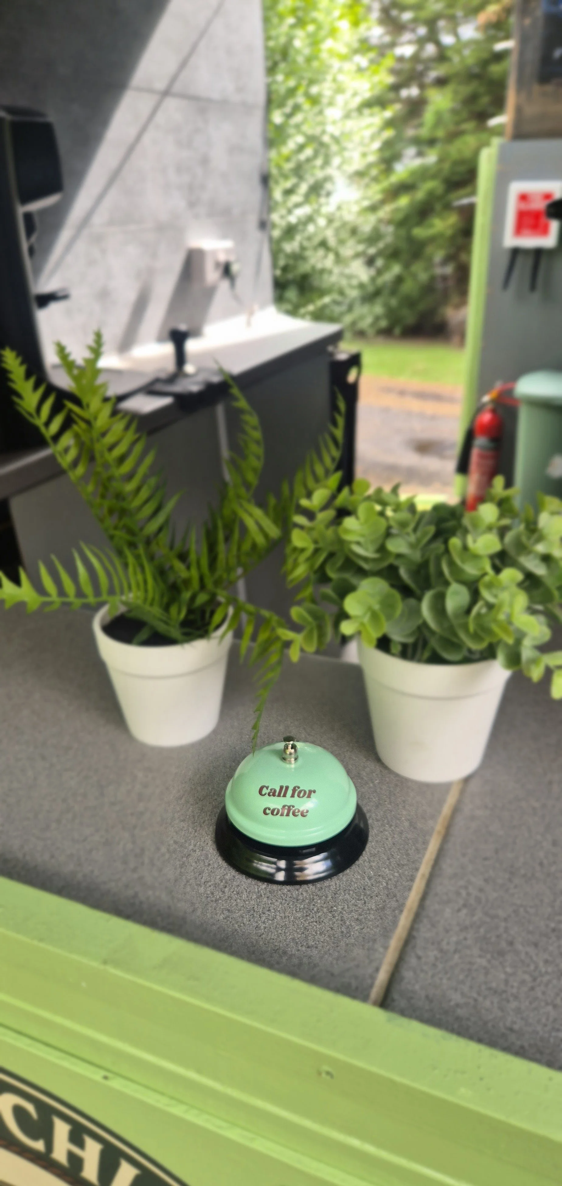 Two potted green plants, a coffee bell with the words "Call for coffee," and a blurred outdoor background.