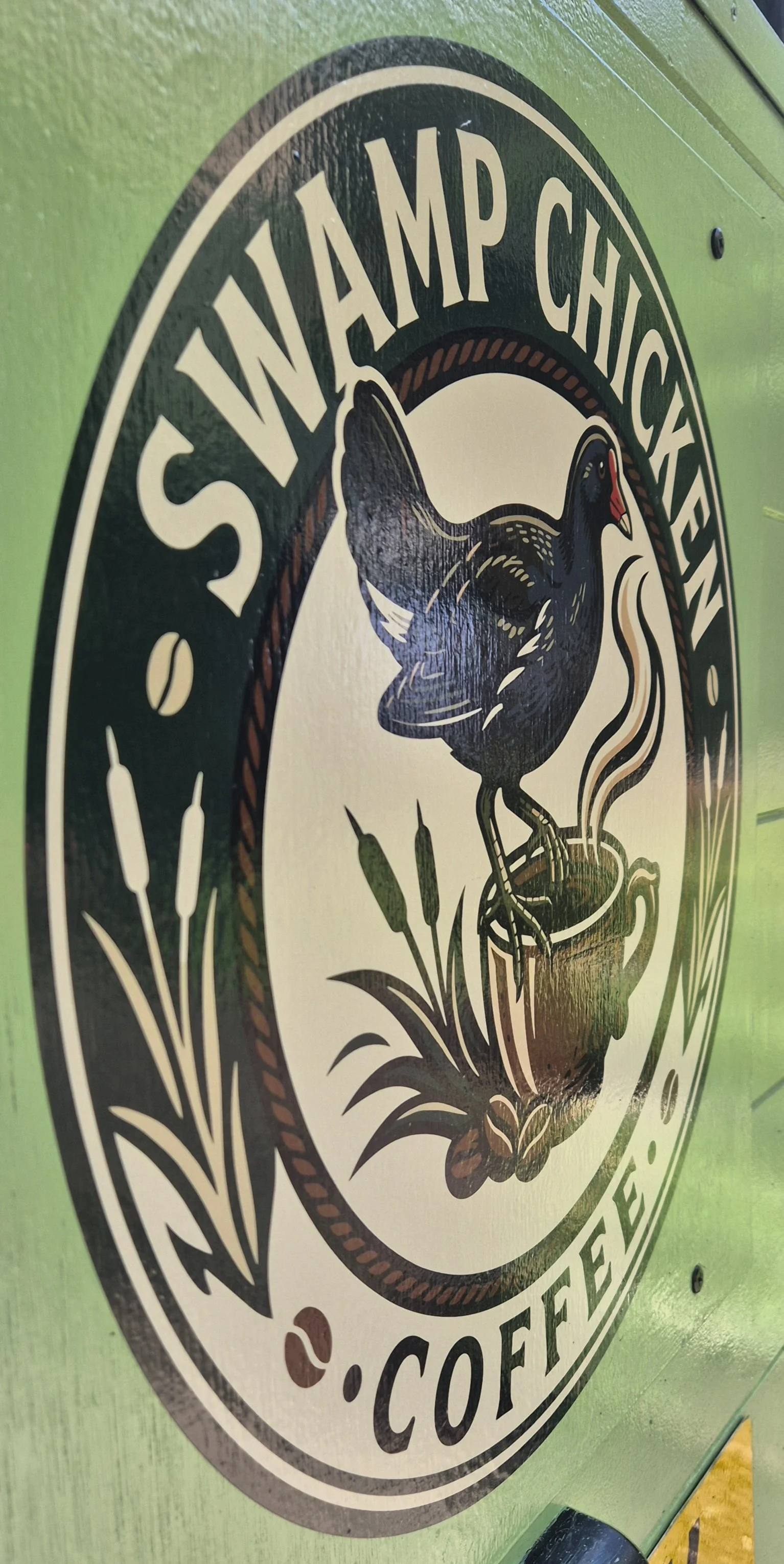 Sign with a black duck sitting on a steaming coffee mug, surrounded by grass and cattails, with the text 'Swamp Chicken Coffee' around the edge.