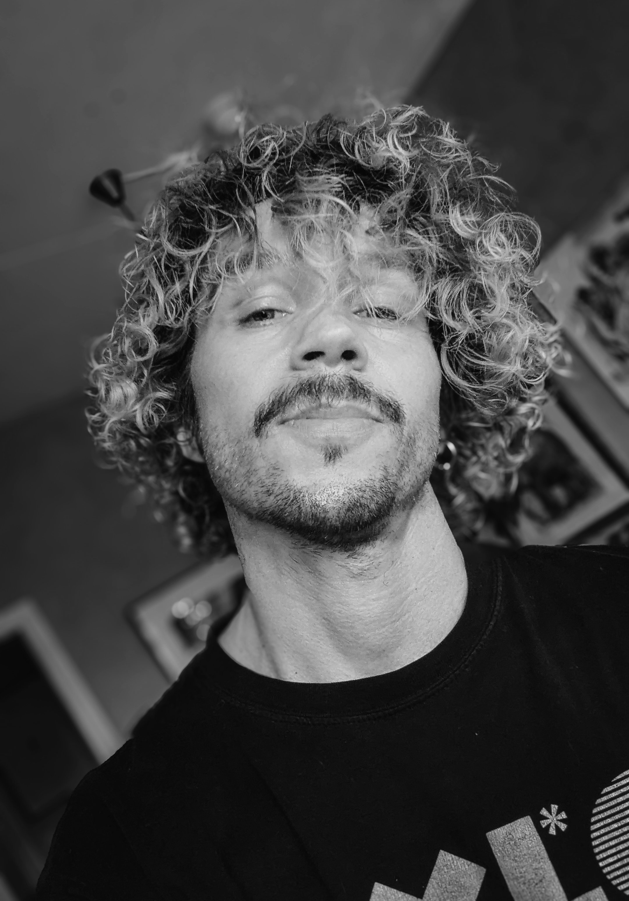 A black and white close-up selfie of a man with curly hair and a mustache, wearing a black T-shirt with a graphic design.