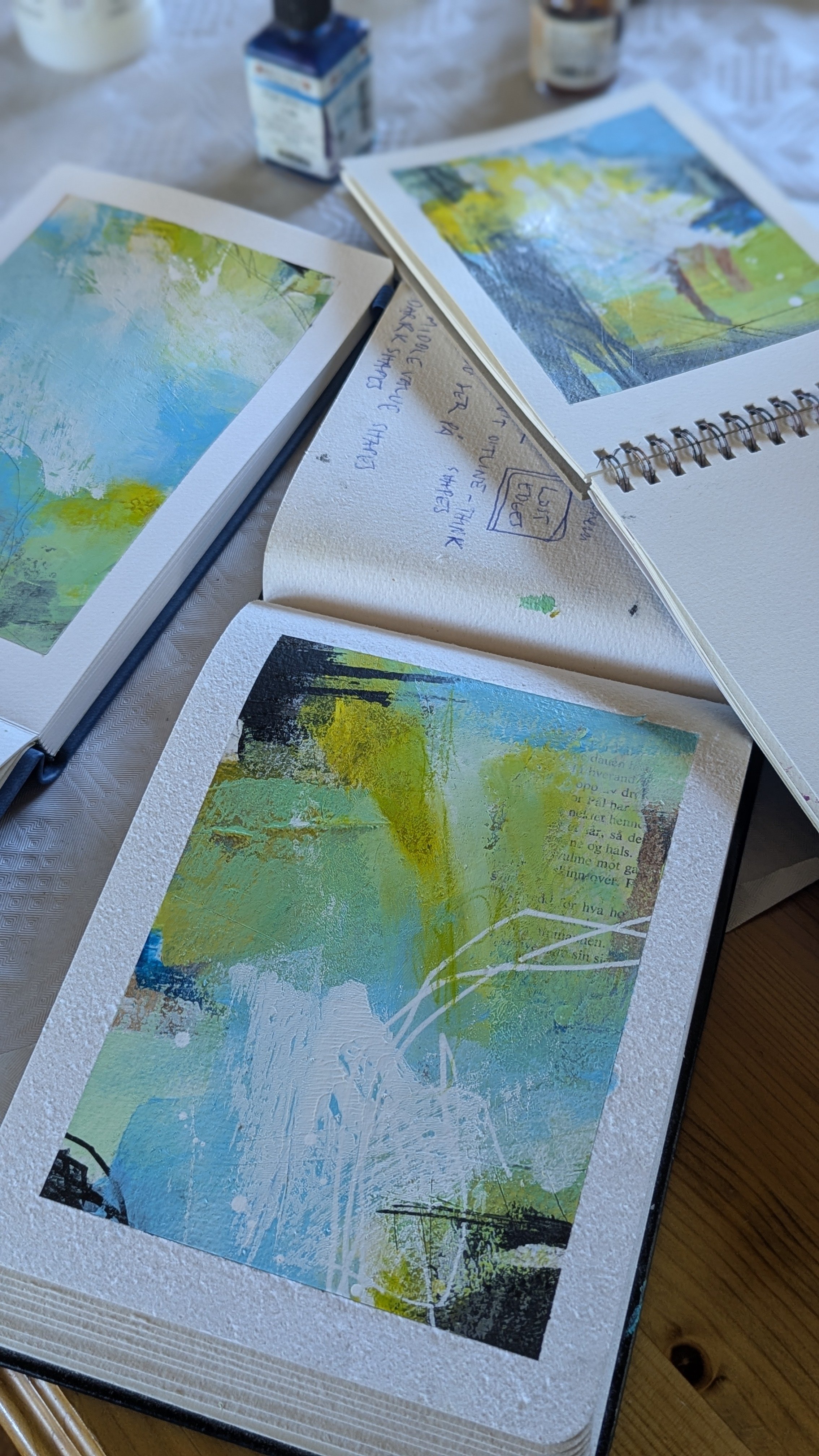 Open art print portfolio with three abstract landscape paintings in shades of green, blue, and yellow, alongside handwritten notes and art supplies on a table.