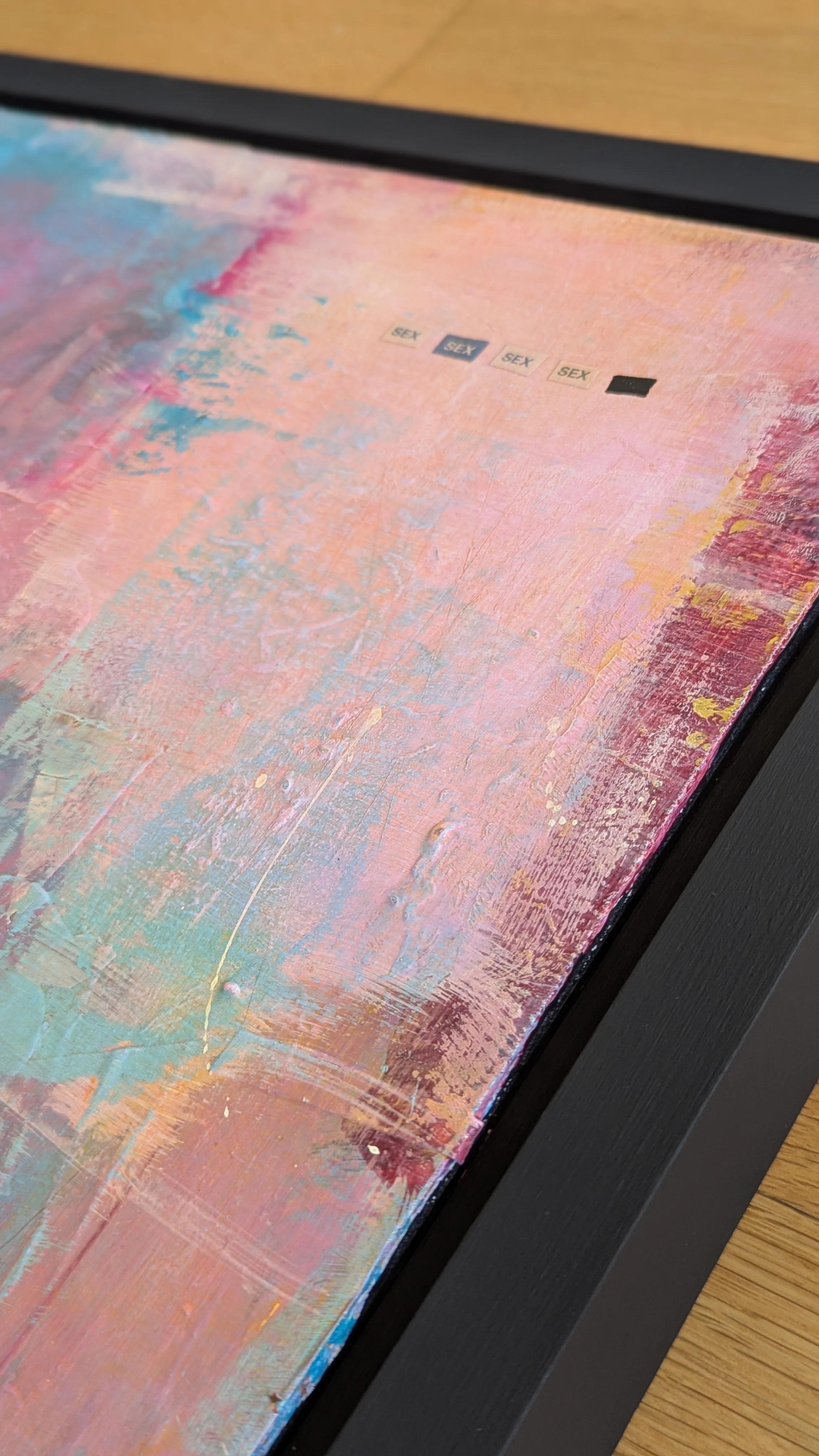 Close-up of a colorful abstract painting with layers of pink, blue, and orange hues, and three small labels that read 'SEX' on the canvas.