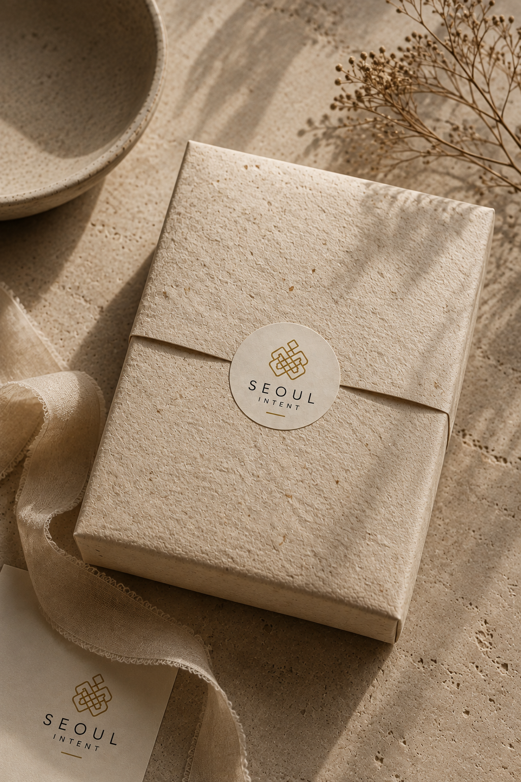 A beige textured gift box with a round sticker labeled 'SEoul Intent' on top, next to a matching ribbon and a piece of fabric, with a ceramic cup and dried flowers nearby.