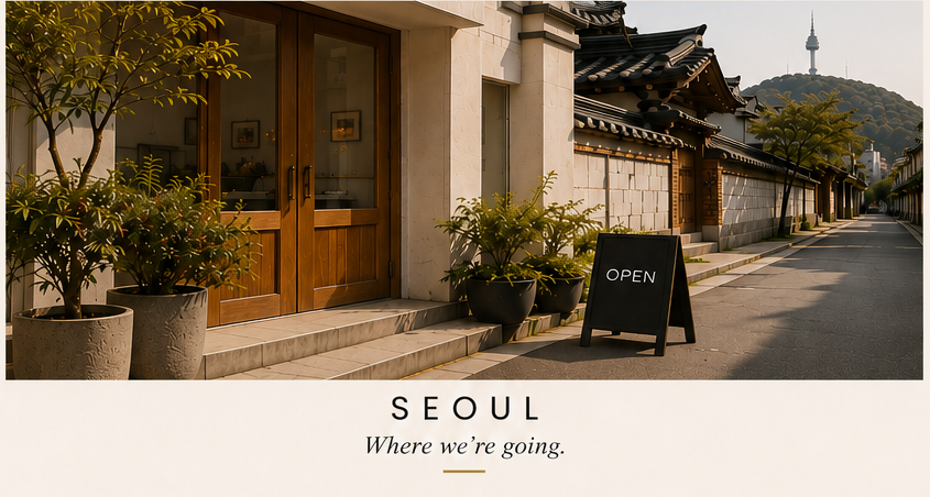 Street view of a traditional Korean building with a wooden door, potted plants outside, and an 'Open' sign. In the background, Namsan Tower is visible on a hill.