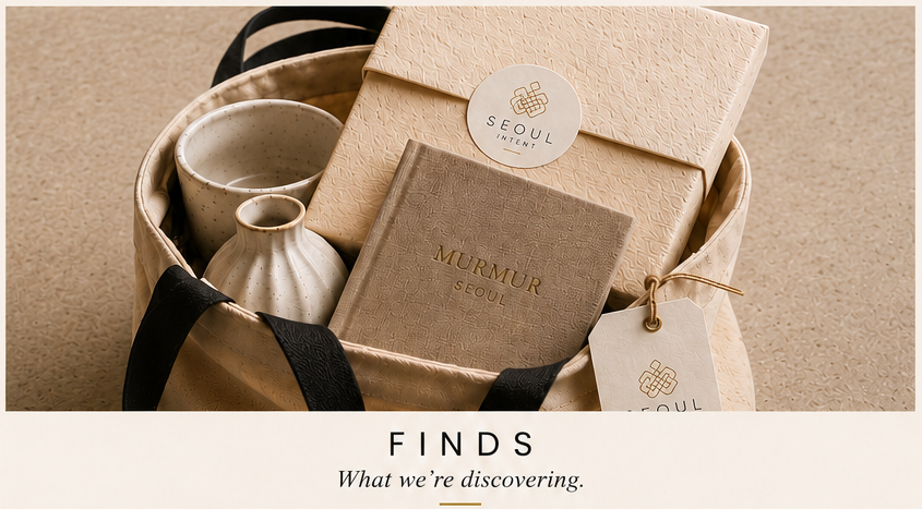 A gift basket containing a beige box with a Seoul Intents logo, a small beige booklet, and two ceramic cups, one larger and one smaller, all set on a neutral surface with black ribbon handles.