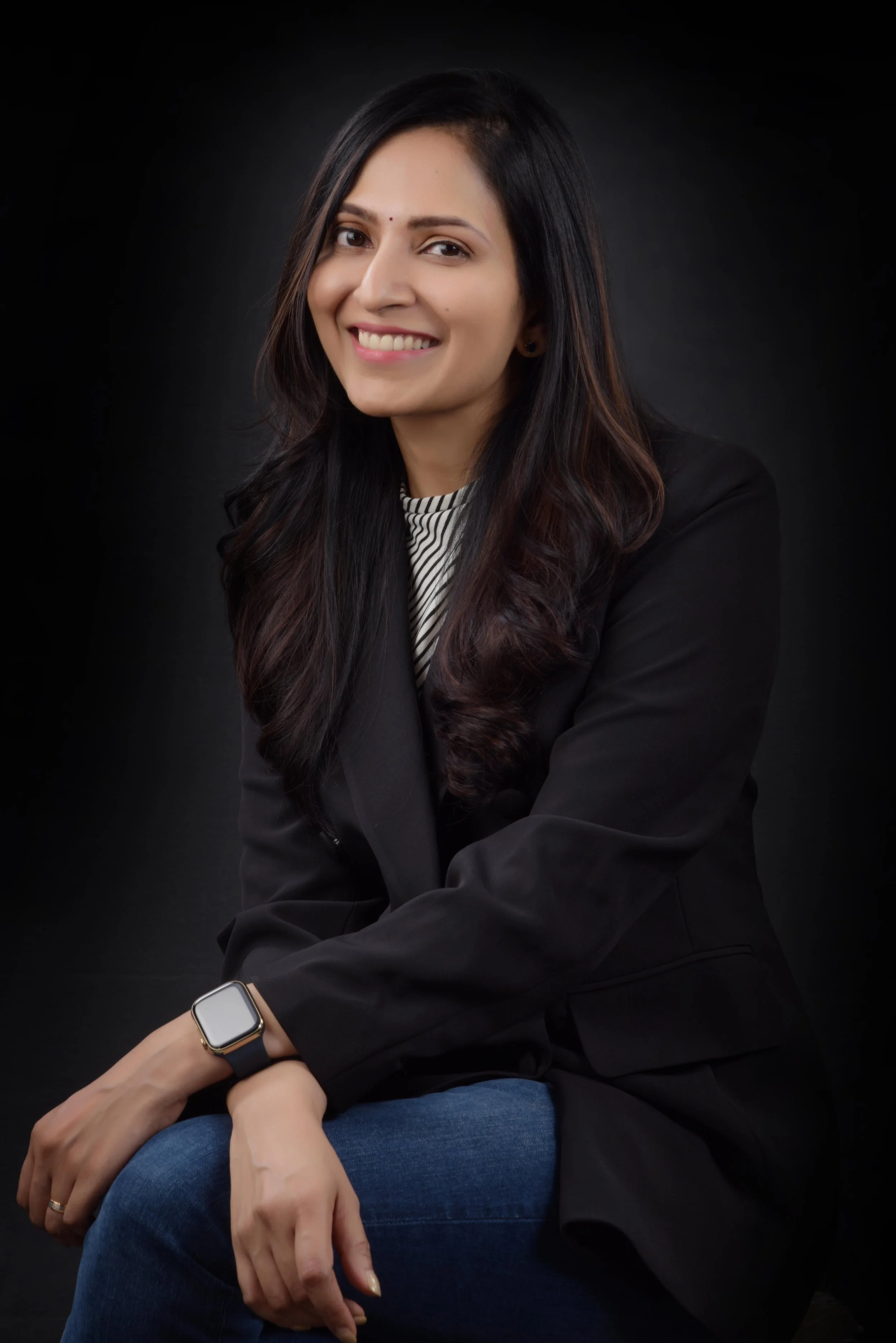 Portrait of a smiling woman with long dark hair wearing a black blazer, striped shirt, and smartwatch, sitting with crossed arms against a black background.
