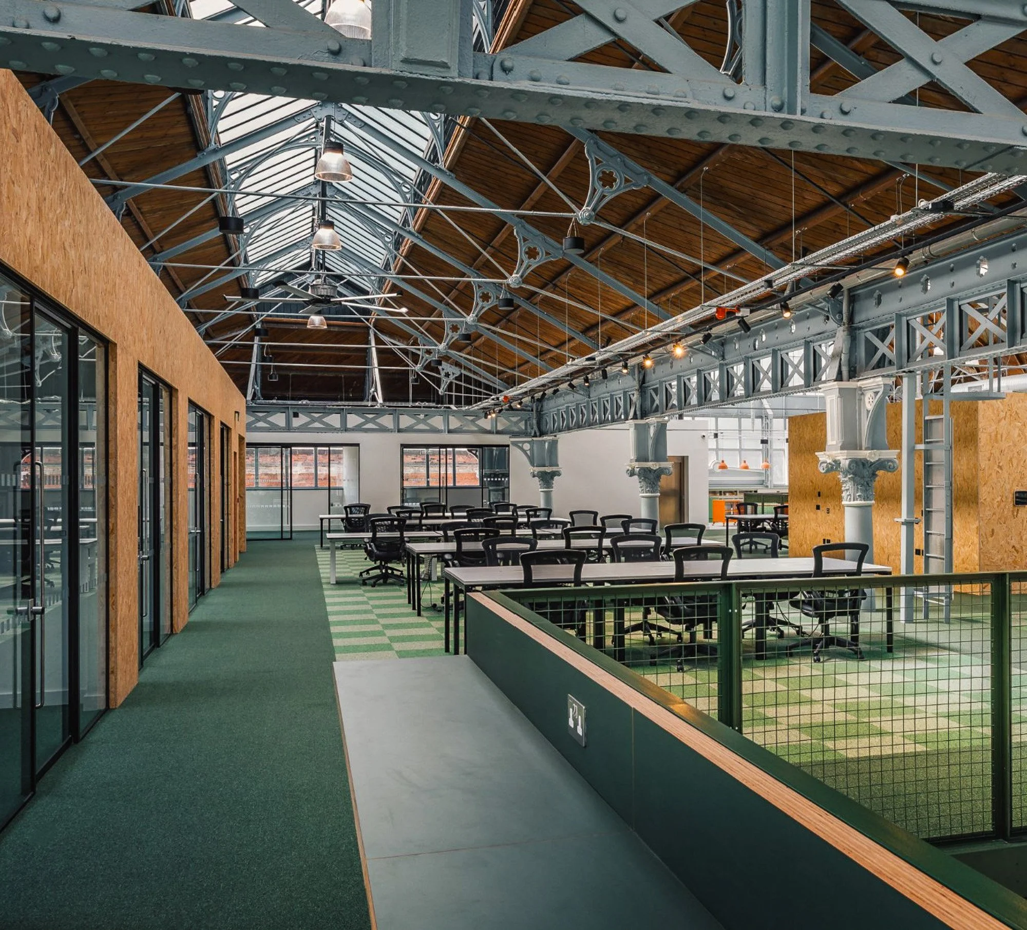 View of an indoor space with a high arched glass and metal roof, exposed beams, and industrial lighting. The space features a green carpeted walkway, glass doors on the left, and tables with chairs inside. The design combines modern industrial and vintage elements.