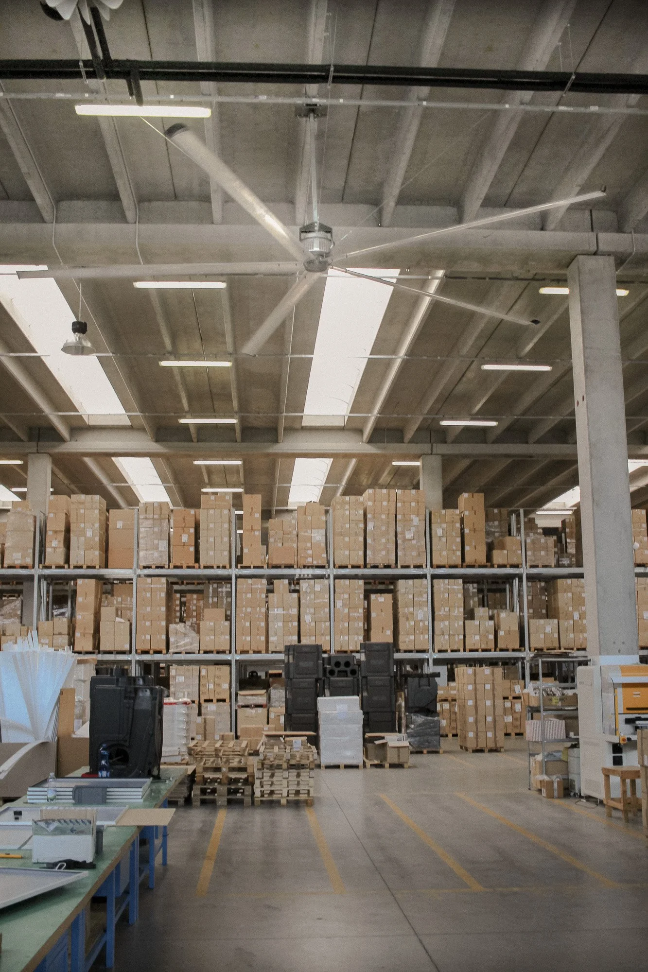 Warehouse with stacked boxes on shelves, a large ceiling fan, and work tables.