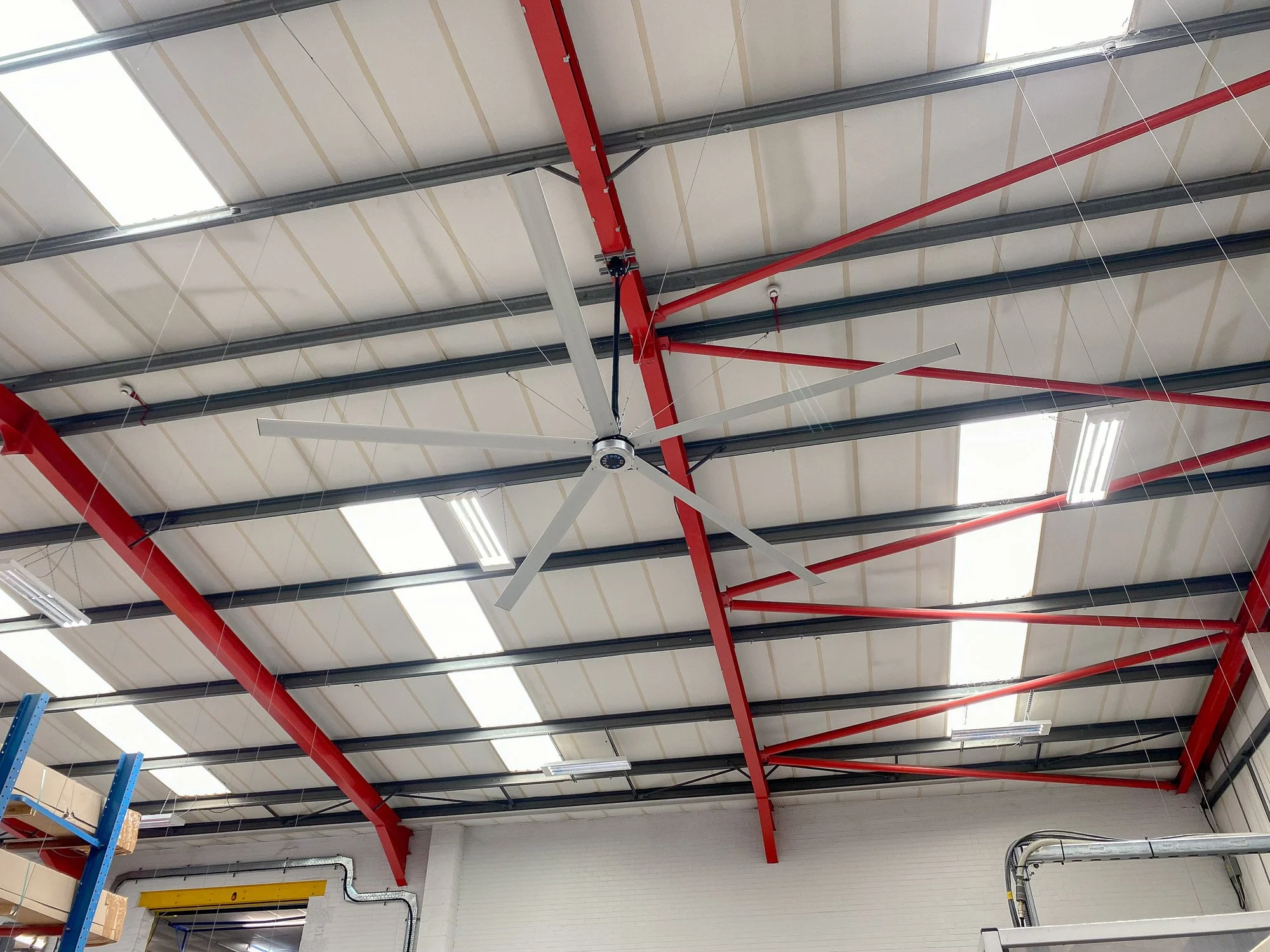 Interior of a warehouse or industrial building ceiling, with exposed metal beams, red support structures, a large industrial fan, and overhead lighting panels.