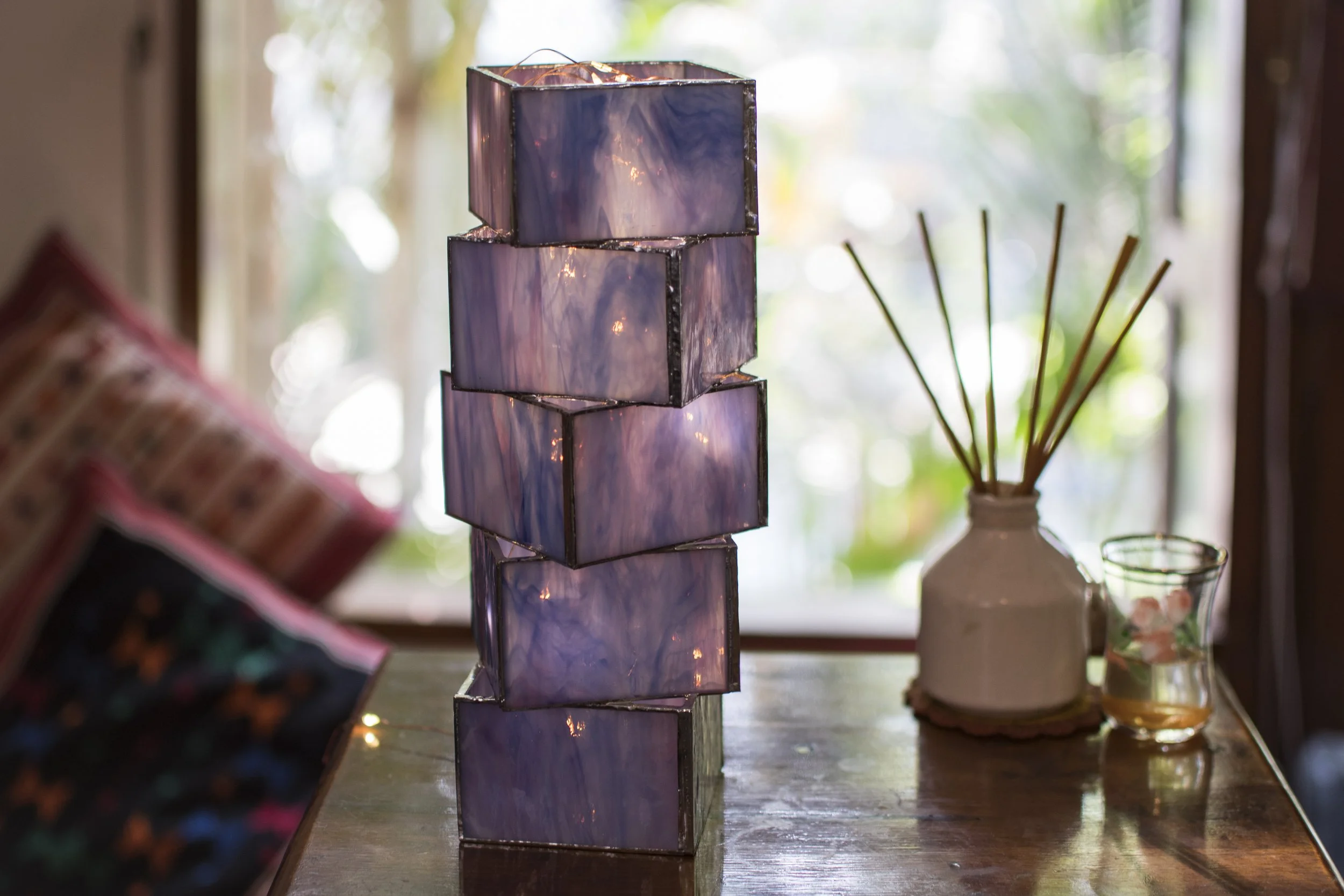 A table with a stack of four hammered purple glass cubes with glowing lights inside, next to a white vase with reed diffuser sticks and a small glass bowl.