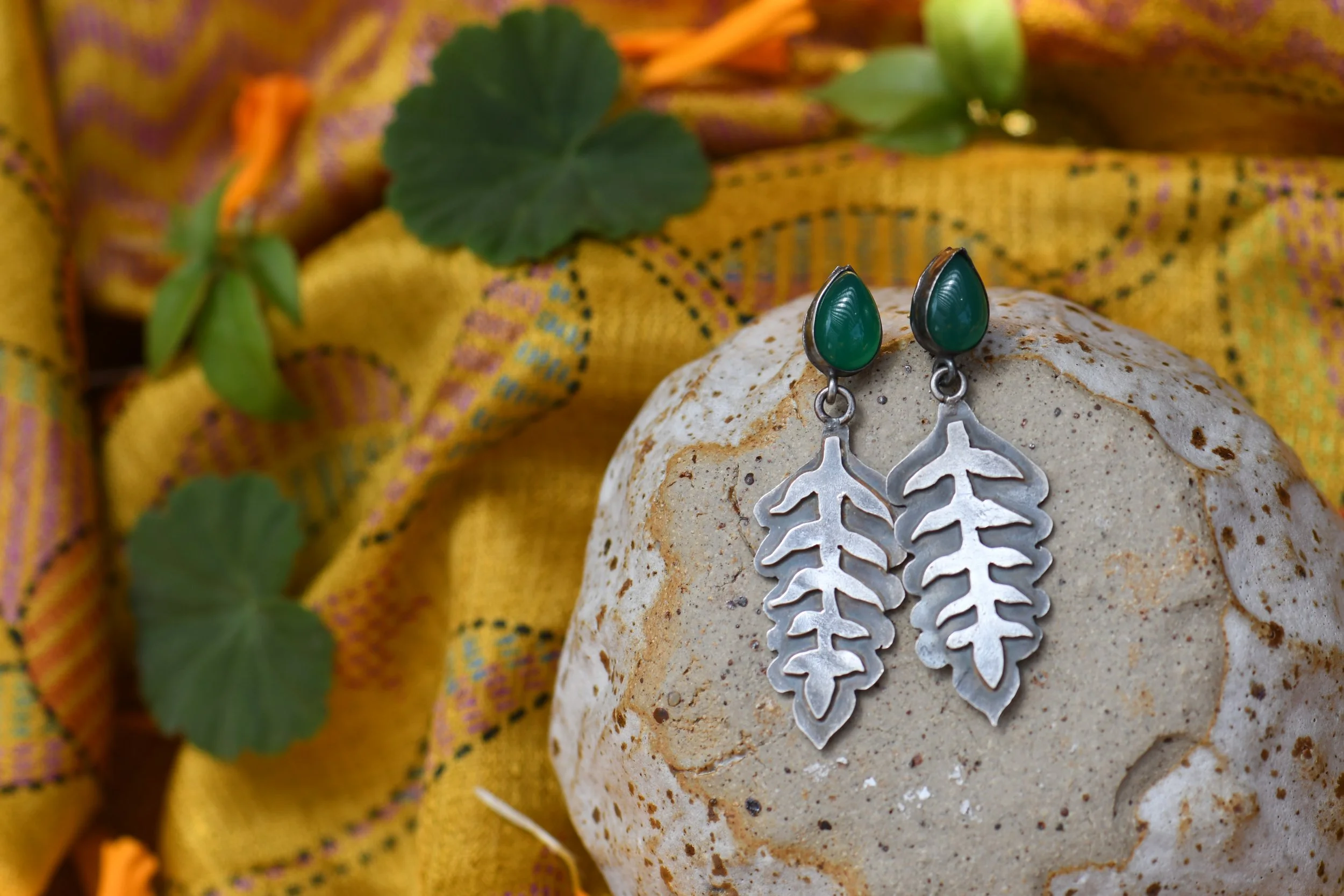 Silver earrings with green stones and leaf-shaped pendants on a textured stone, with yellow patterned fabric and green leaves in the background.