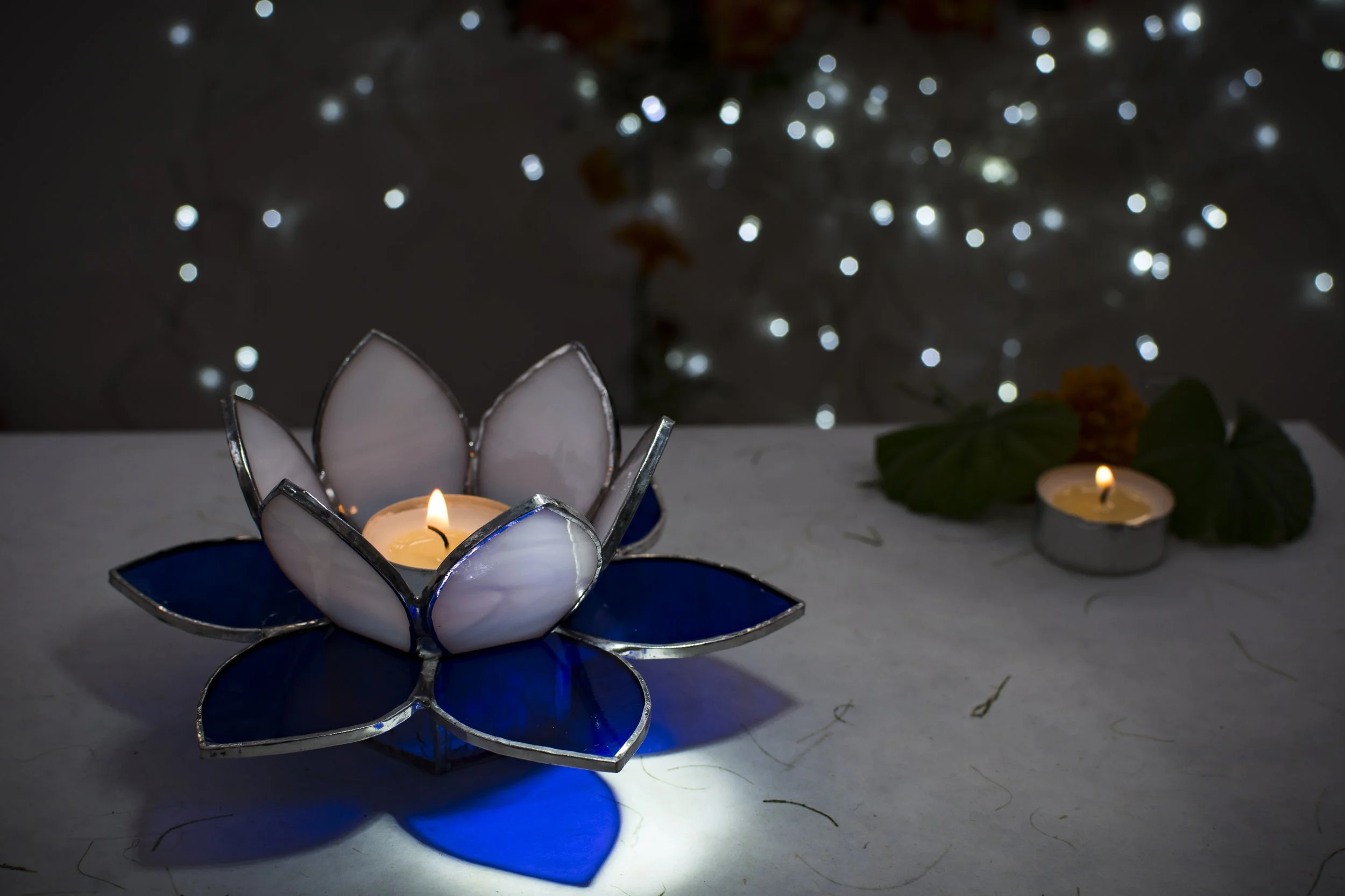 A decorative candle holder shaped like a flower, with a lit candle in the center, placed on a table. In the background, there are blurred string lights and another small candle with green leaves and orange flowers nearby.