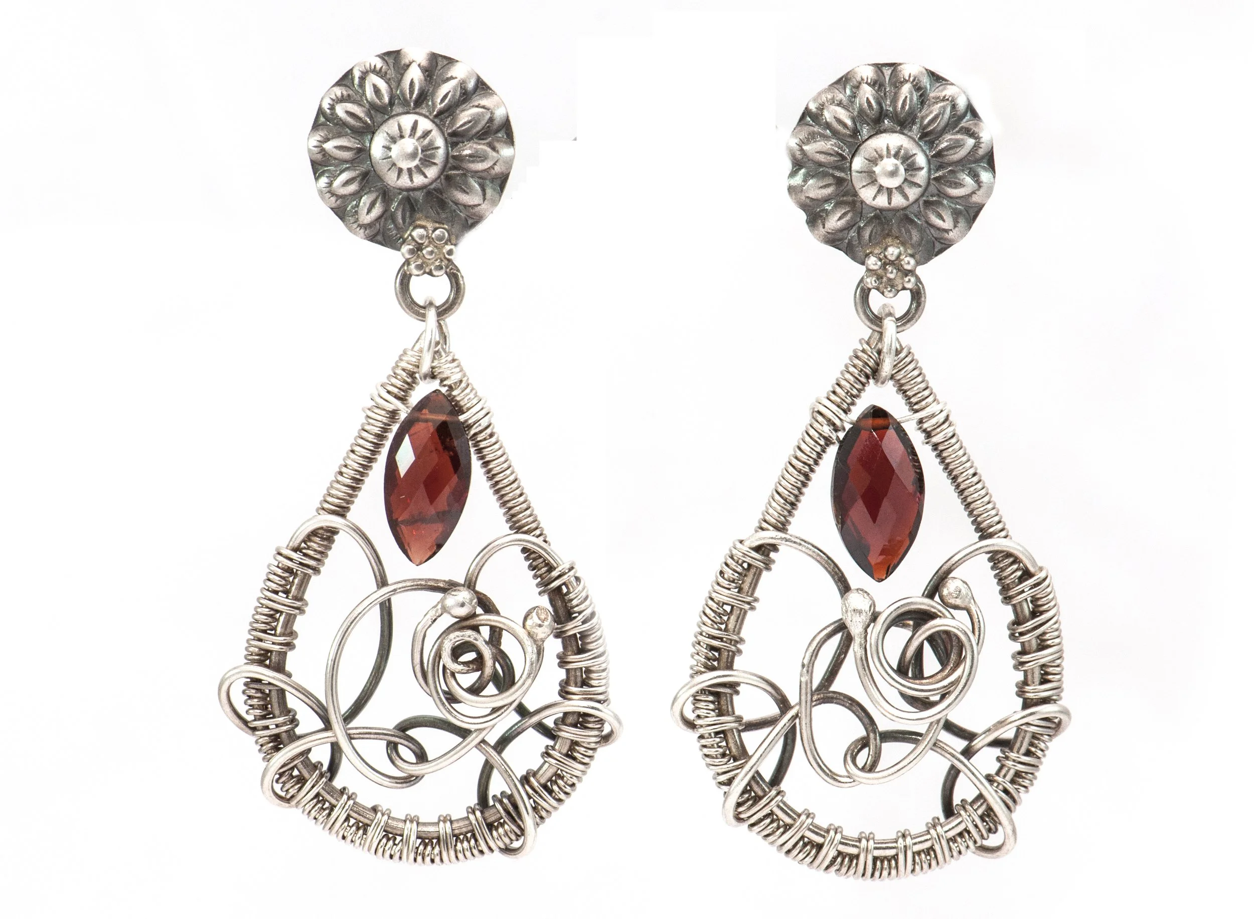 Pair of earrings made of silver wire, featuring a floral design at the top and a red teardrop-shaped gemstone dangling from the bottom.