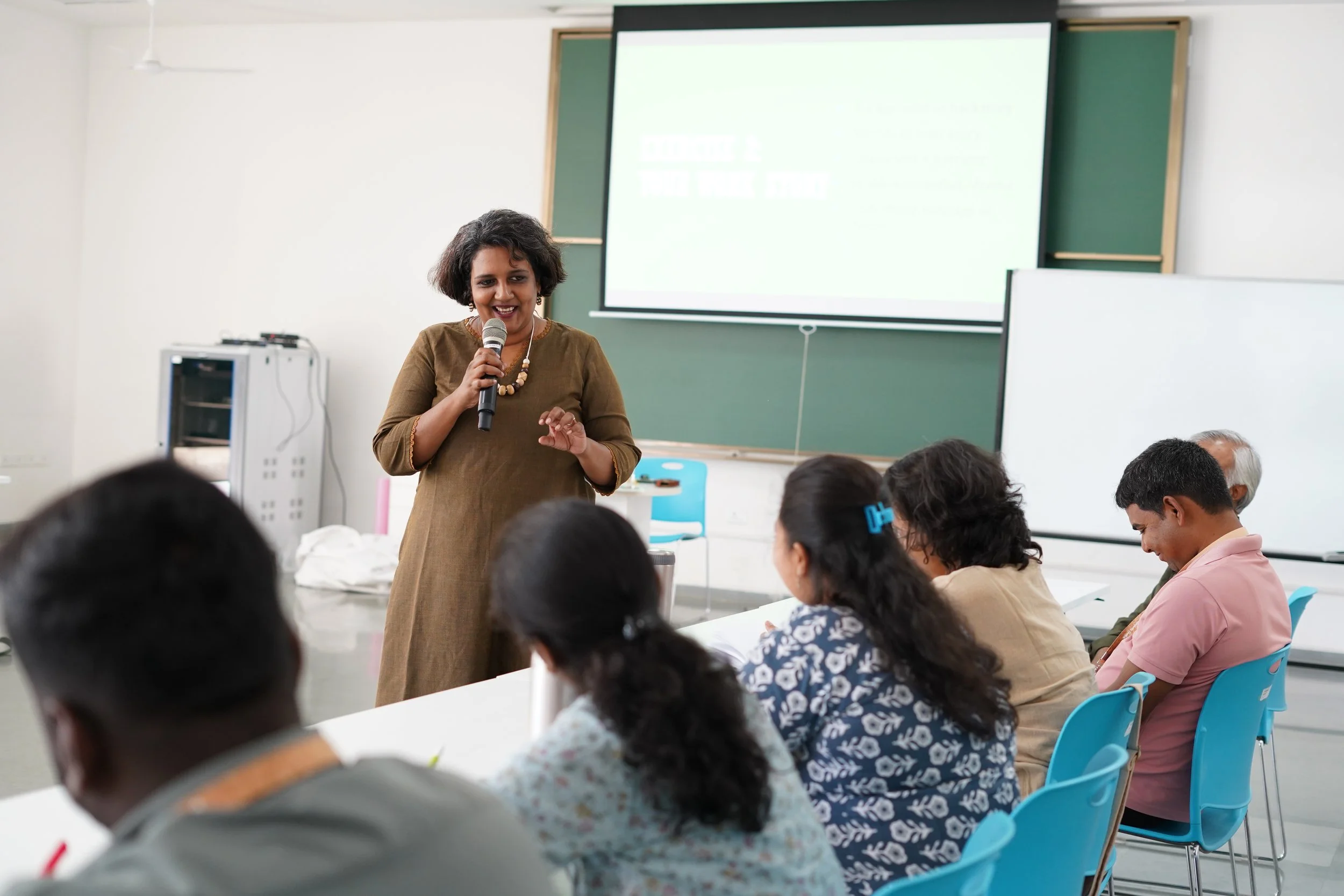 A woman is giving a presentation to a group of people in a classroom. She is holding a microphone and standing near a chalkboard and a projector screen. The audience includes men and women seated at tables, listening attentively.