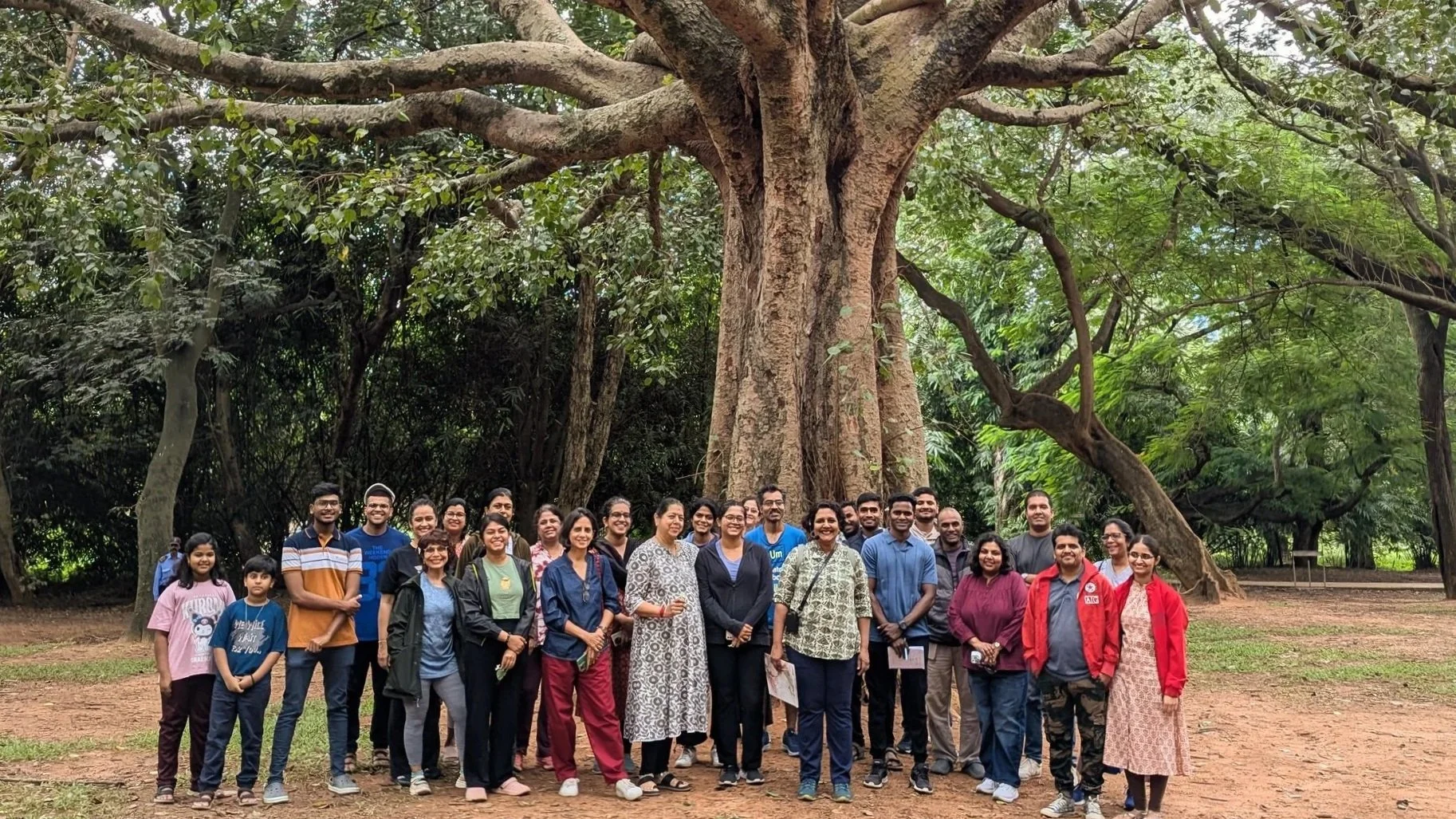 A group of 21 people, including men, women, and children of South Asian descent, standing outdoors in front of a large tree with sprawling branches in a forested area, smiling at the camera.