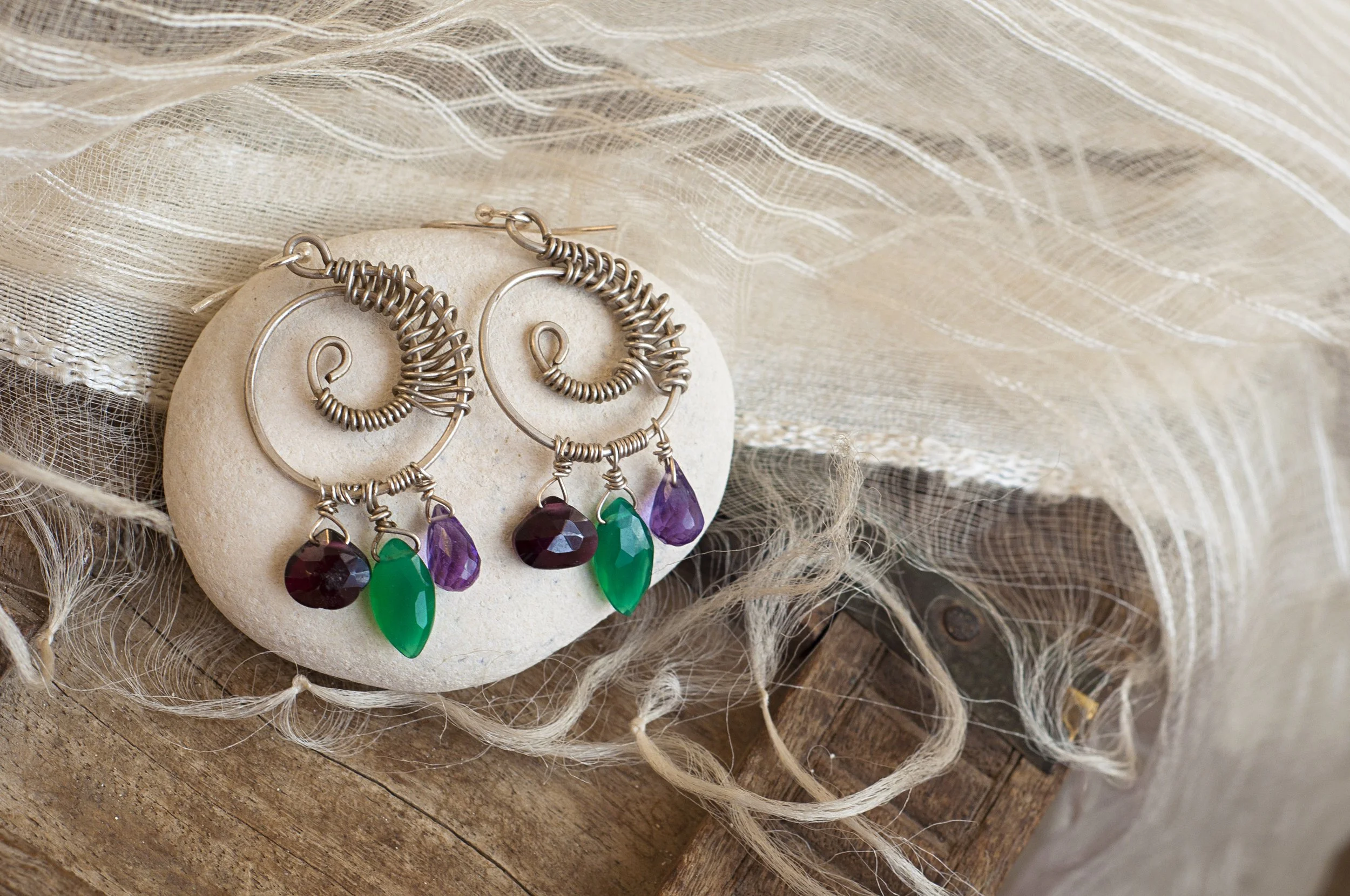 Silver wire earrings with purple and green teardrop-shaped gemstones, displayed on a beige circular card, surrounded by textured cream fabric and a torn burlap cloth.