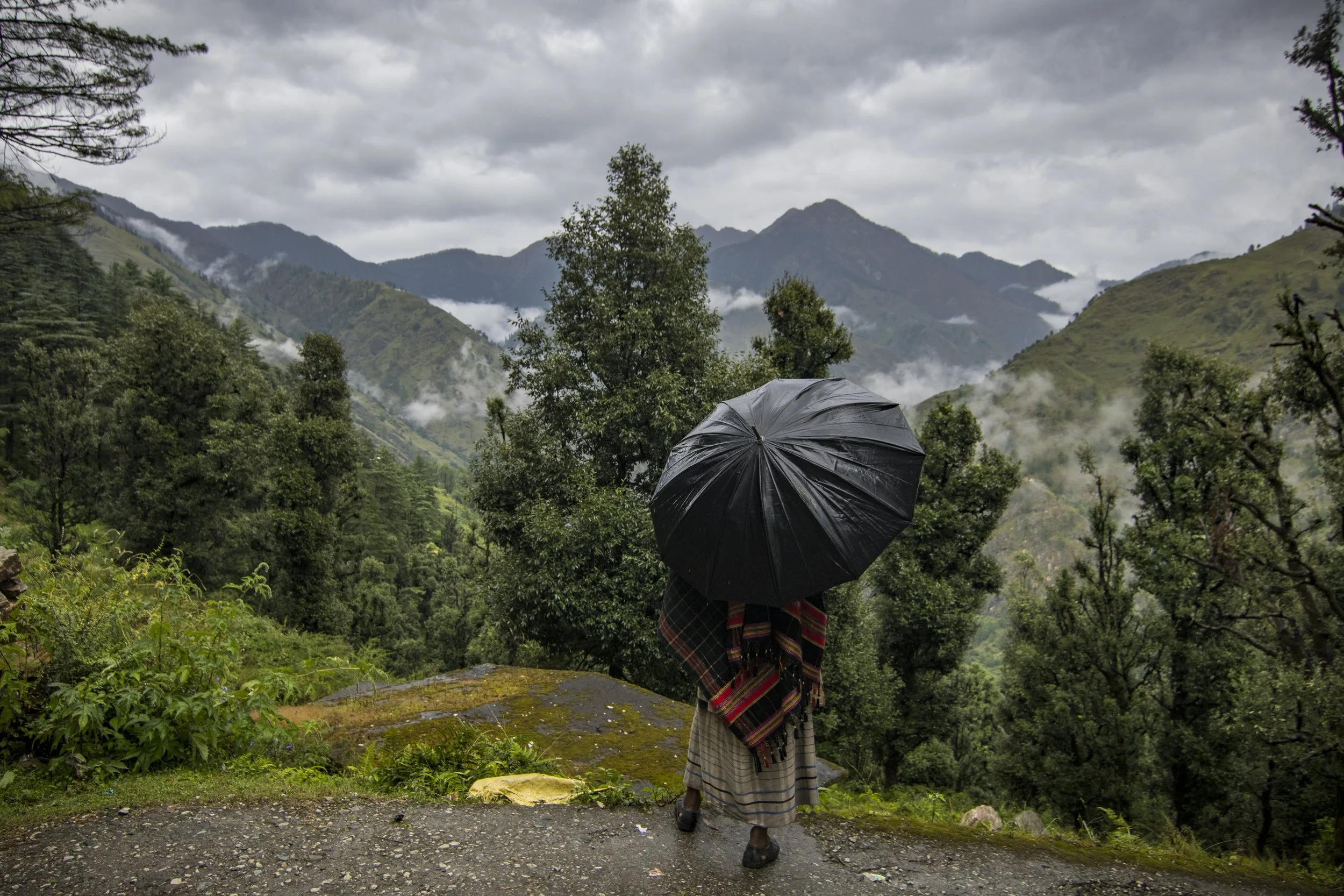 Person wearing traditional clothing holding a black umbrella and overlooking a lush green mountainous landscape with cloudy sky.