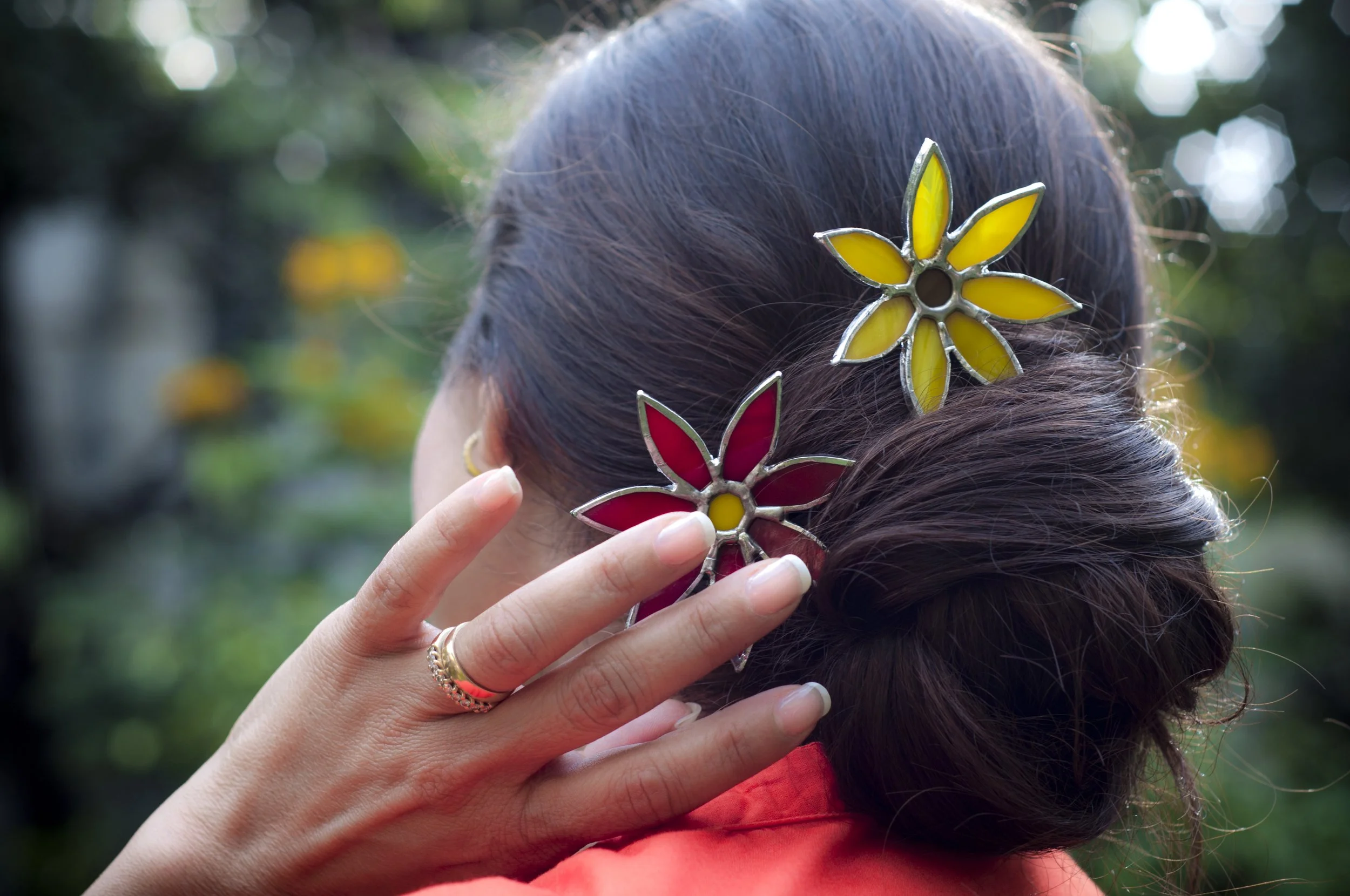 A person adjusting a yellow and red flower-shaped hair clip in their dark brown hair outdoors, with green leafy background