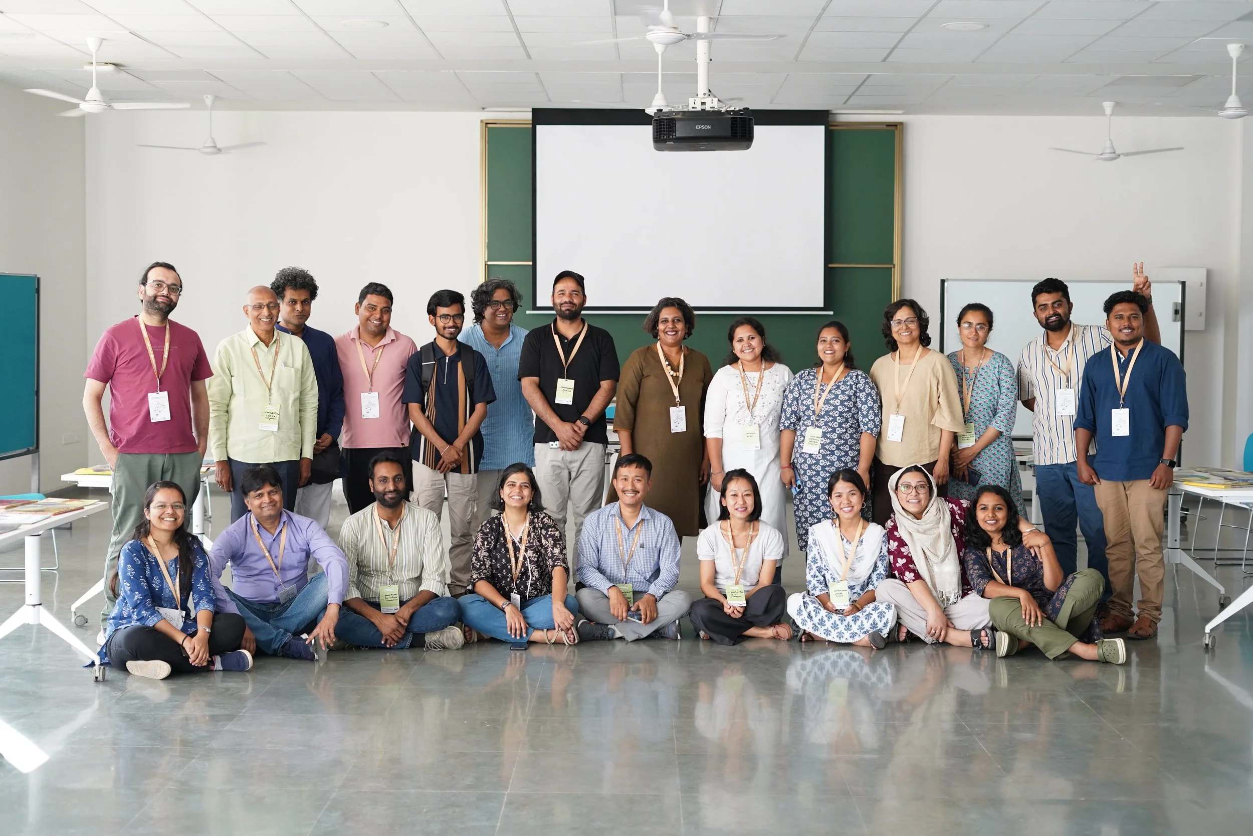 Group photo of diverse people in a classroom or conference room, smiling and posing for a picture, with a presentation screen and whiteboard in the background.
