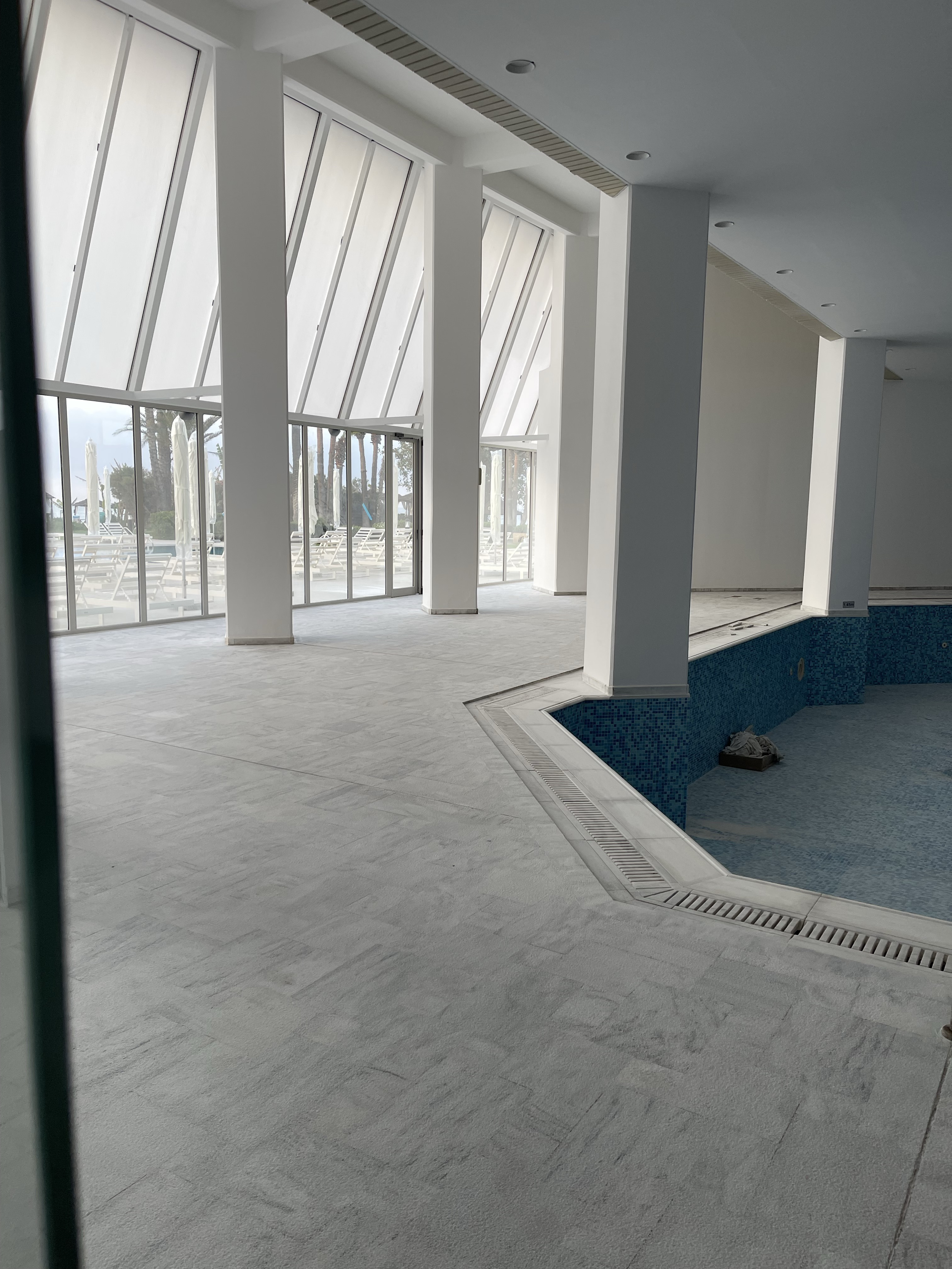Empty indoor pool area with large windows and lounge chairs outside.