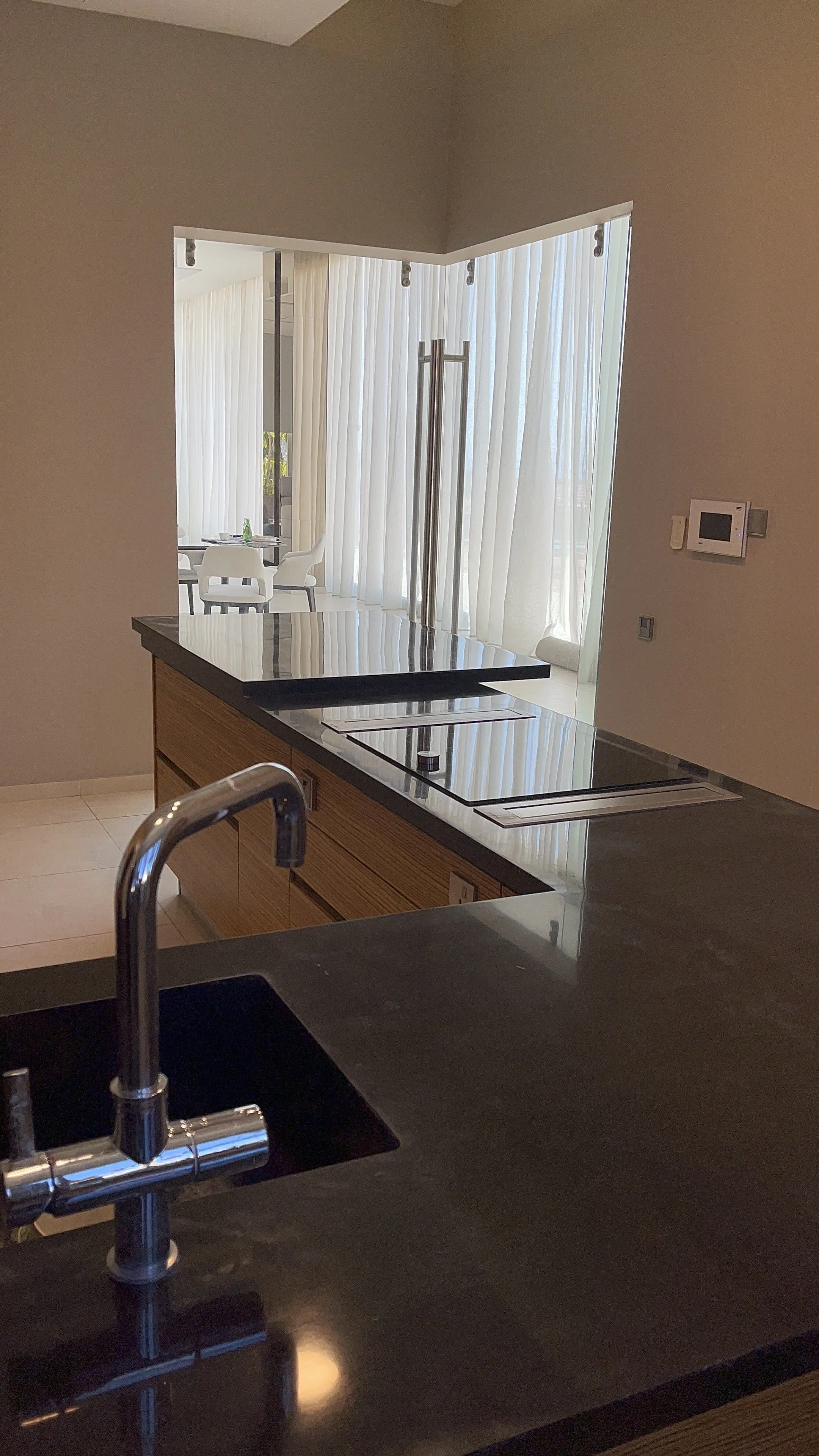 View of a modern kitchen with a black countertop and stainless steel sink, looking out into a room with white chairs and tall white curtains.
