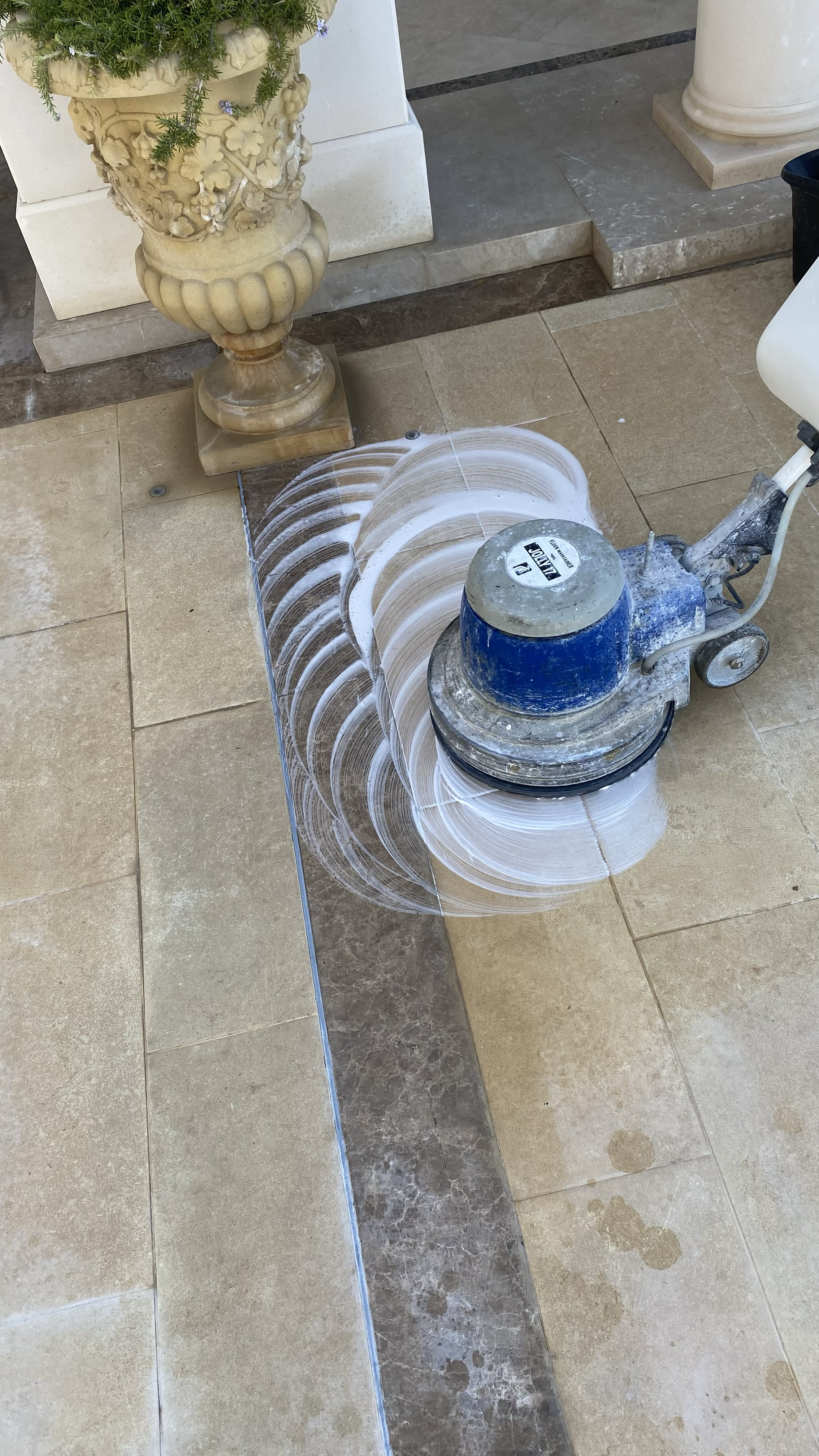 A floor cleaning machine is working on a tiled floor, creating a circular pattern with its cleaning motion. There are decorative pillars and a large ornate flowerpot with greenery and flowers in the background.