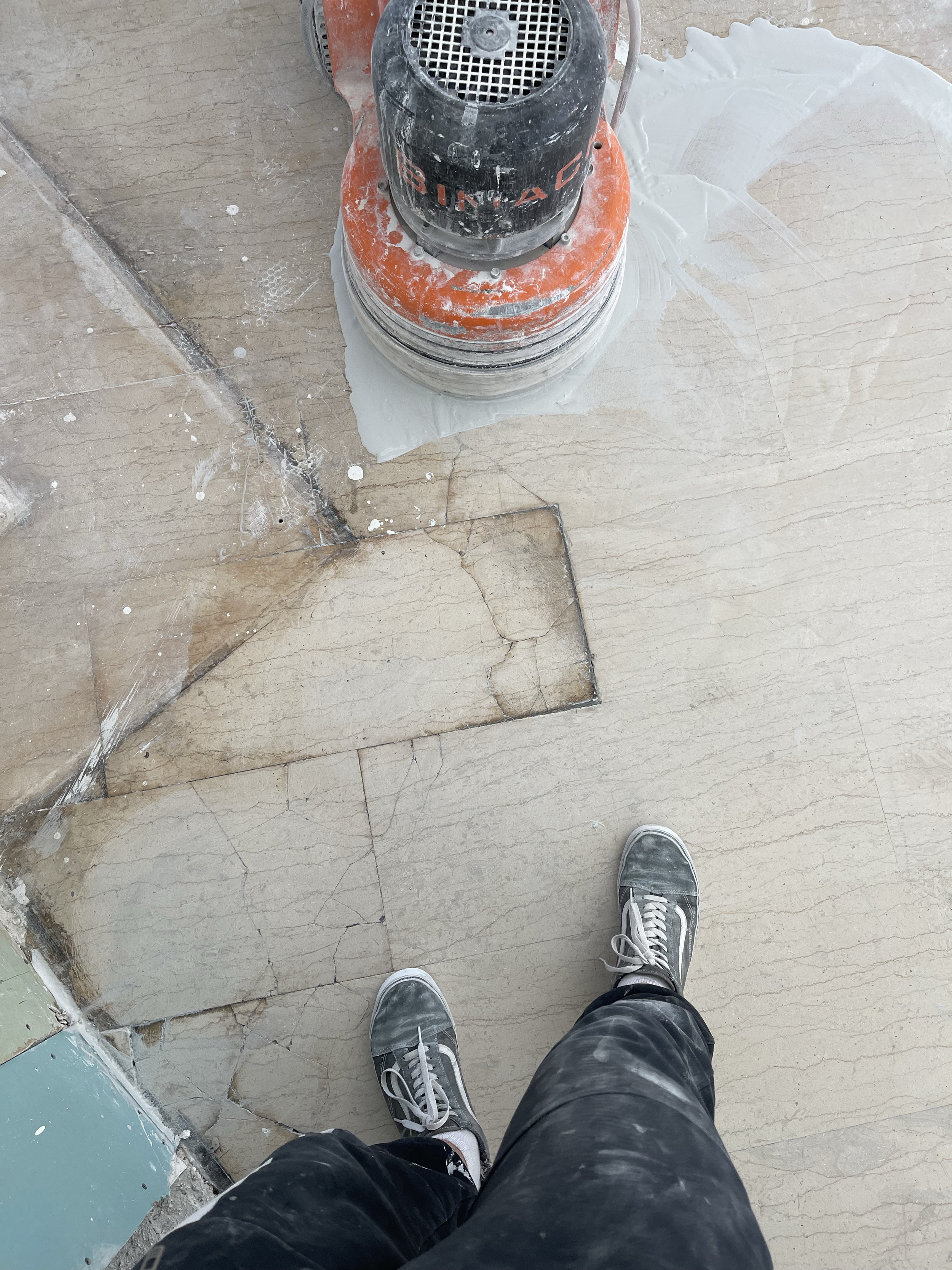 A person standing on a construction site wearing black sneakers and black pants. The floor is under renovation with some tiles removed, exposing a concrete surface underneath. A construction dust extractor or polishing machine is seen on the floor, a