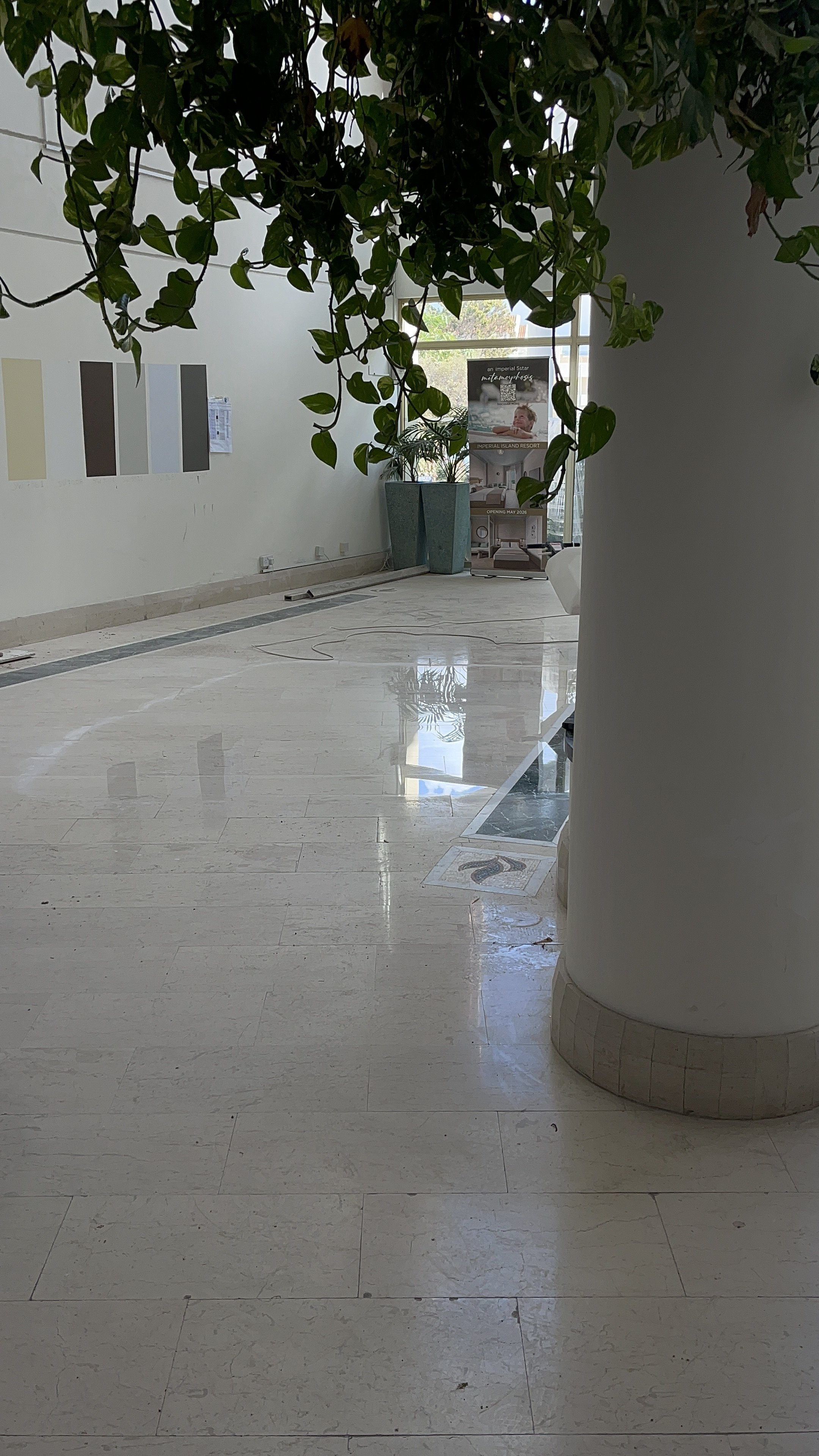 Interior of a building with a tiled marble floor, green plants, and a promotional stand near a large window.