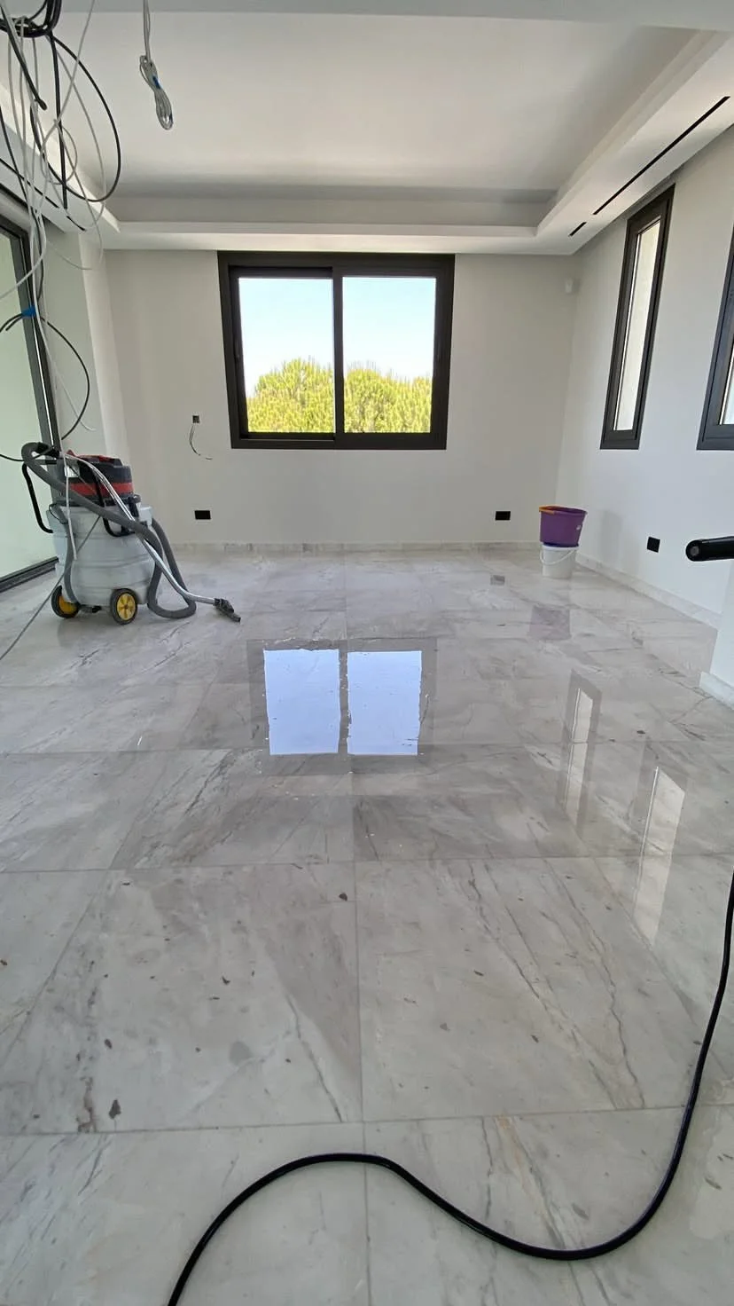 Empty room with marble floor, large windows showing trees outside, construction tools and wiring on the left side.