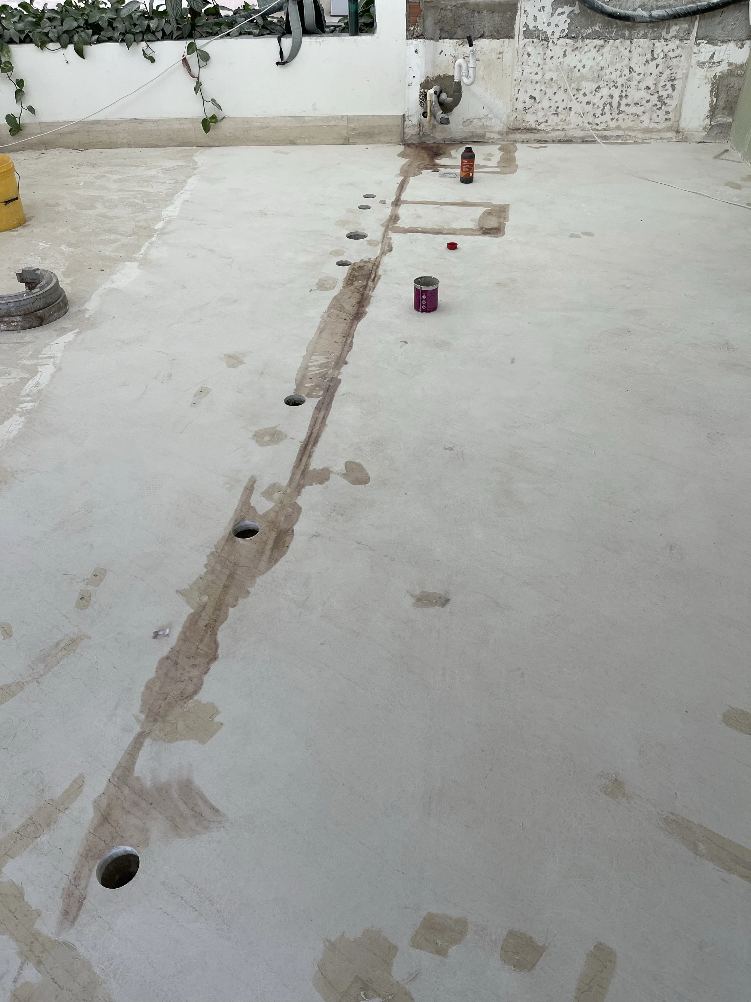 Construction site with a newly poured concrete floor, multiple drainage holes, and construction tools and materials scattered around.