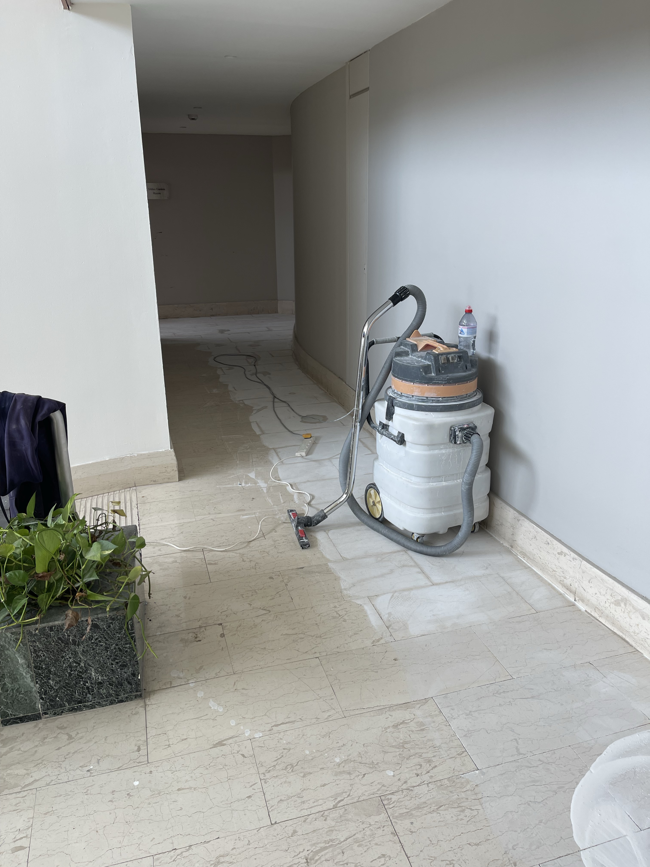 A hallway with beige polished marble tiles and a white wall. There is a potted plant on the left and a cleaning vacuum with a water bottle on top on the right side of the hallway. The ceiling is plain and white.