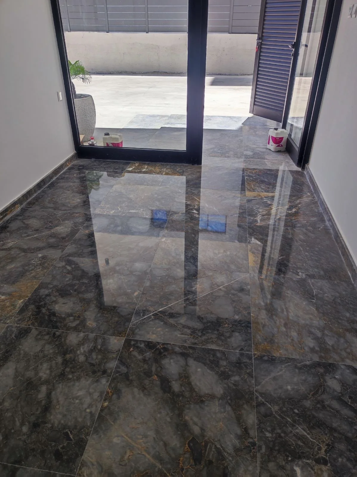 Interior view of a room with shiny dark marble floor tiles, open glass door leading outside, with a potted plant and containers near the door.