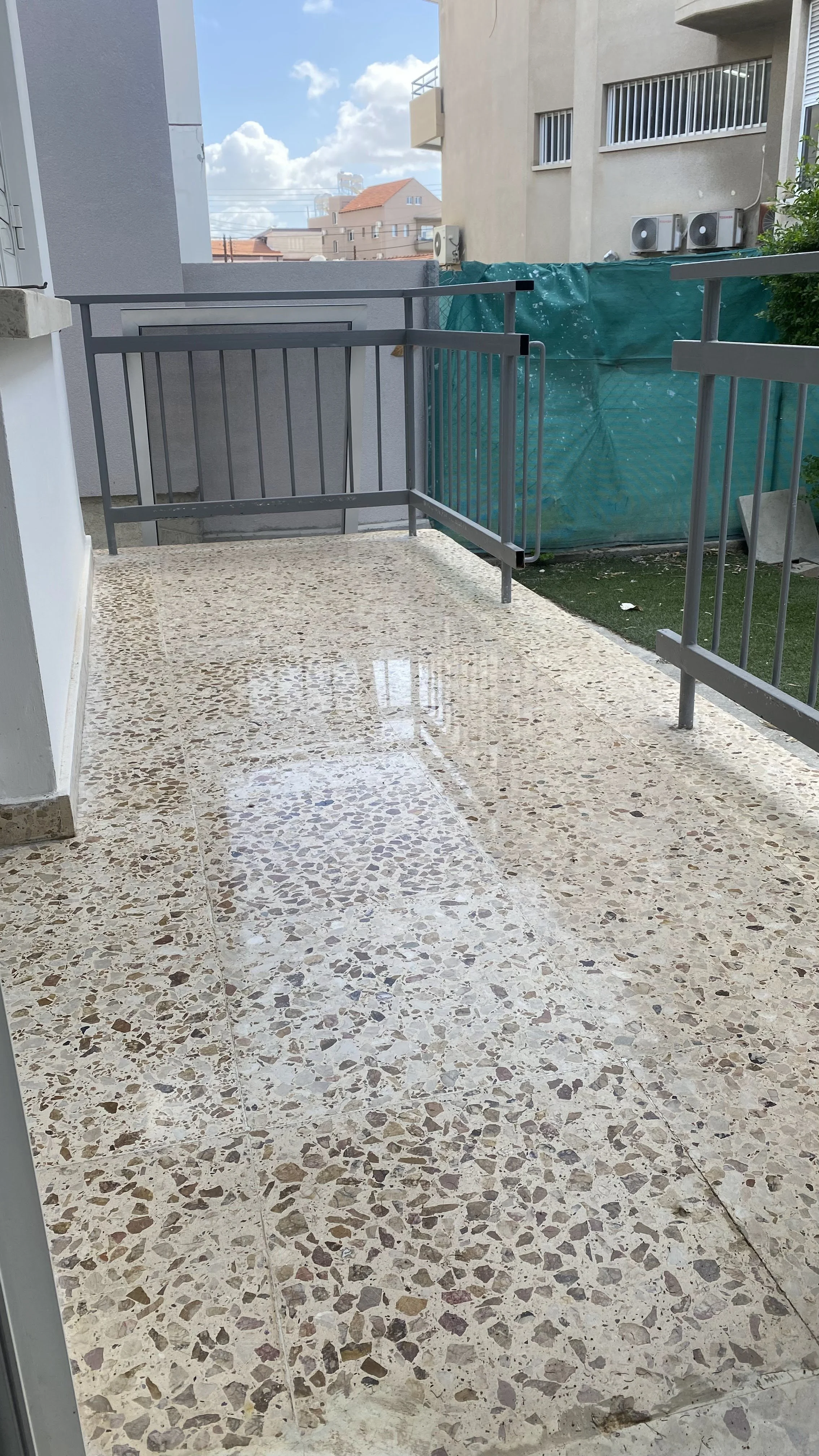 A small balcony with beige terrazzo flooring, gray metal railings, and a view of neighboring buildings under a partly cloudy sky.