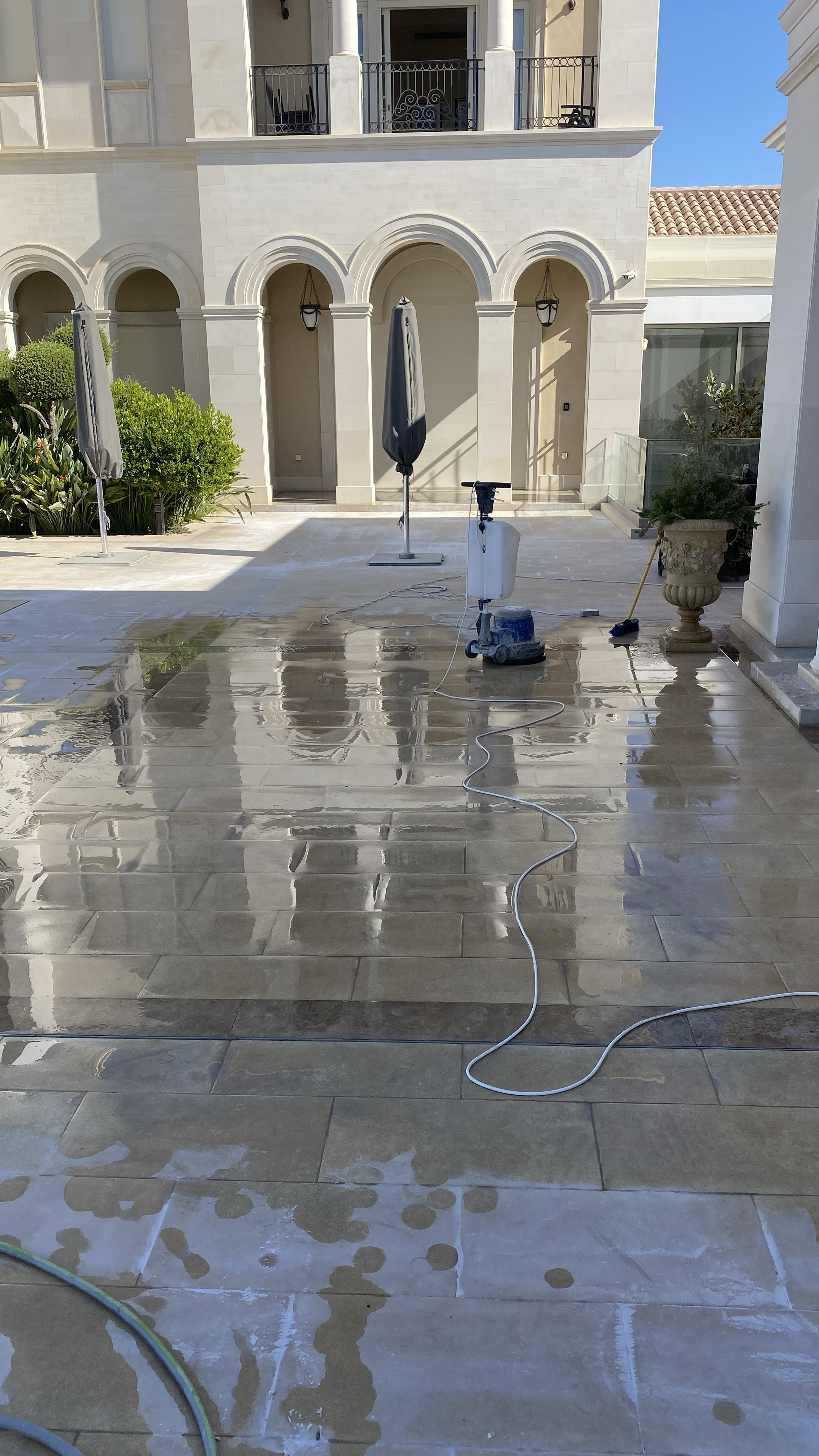 An outdoor patio area being cleaned with a pressure washer, showing wet tiled flooring, a closed umbrella, and a residential building with arched doorways and balconies.