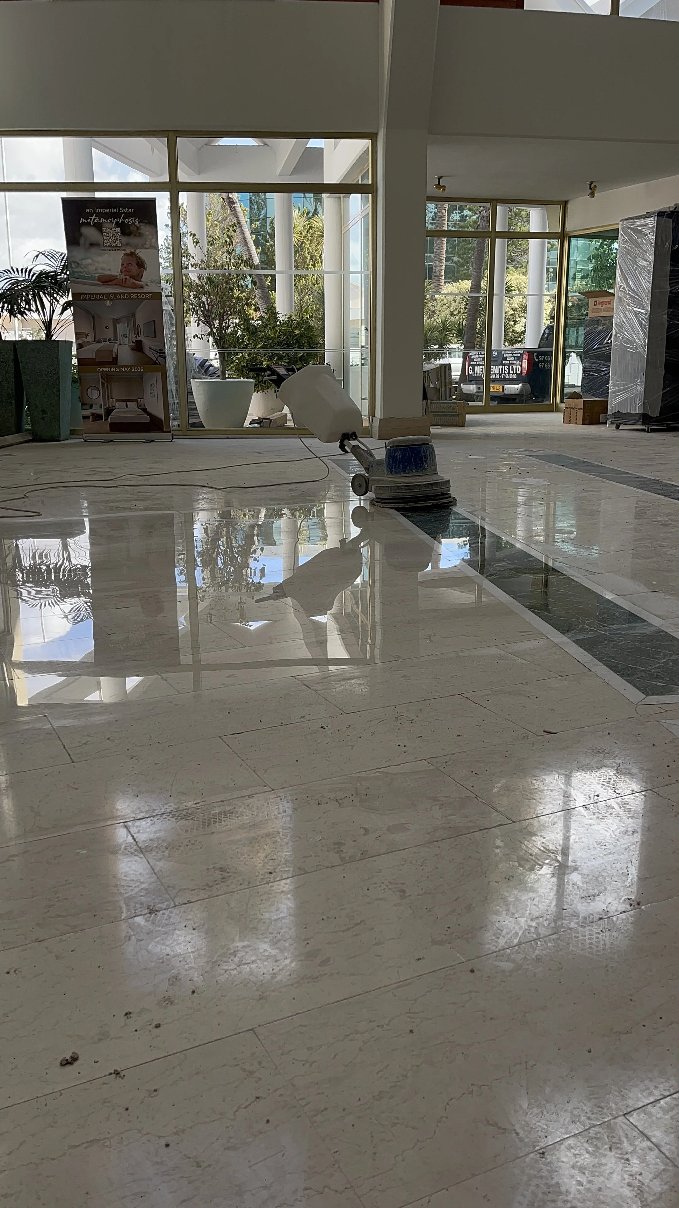 A clean, shiny marble floor inside a building with glass walls and windows showing trees and a vehicle outside. There is a floor cleaning machine with a white head and blue handle in the center.