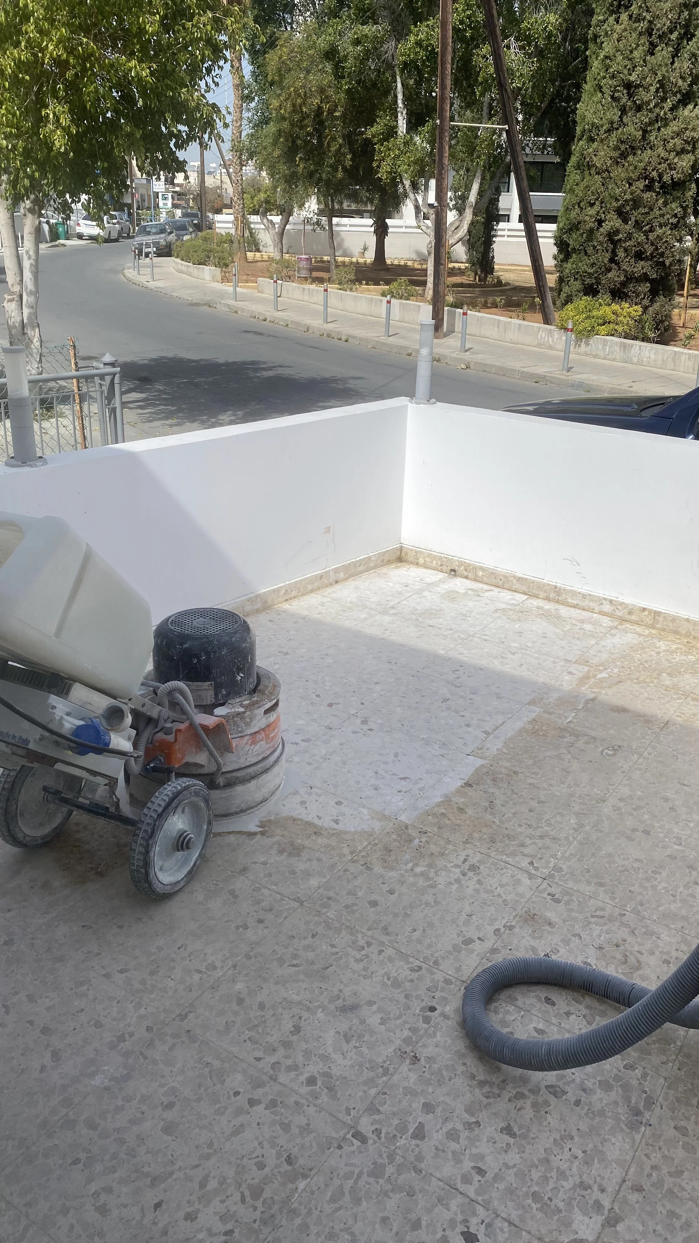 View of an outdoor patio area with construction or renovation work in progress, showing a floor grinding machine, a hose, and a white wall in the foreground, with trees, a street, and parked cars in the background.