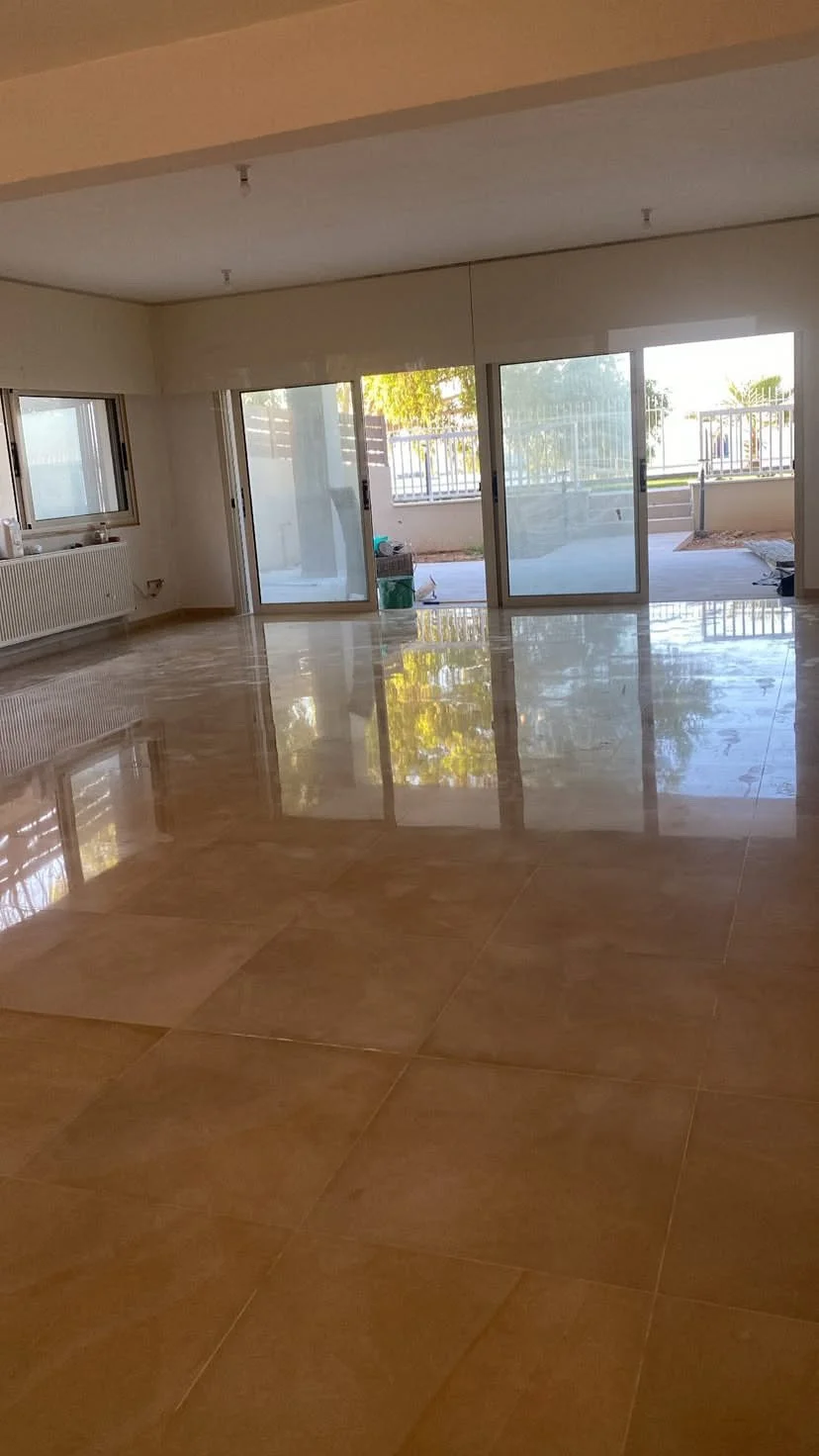 Empty room with shiny beige tiled floor, large glass sliding doors leading to an outdoor patio, with some construction materials outside, and a window on the left wall.