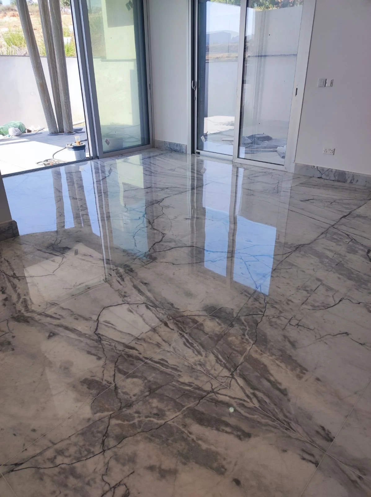 A room with a highly polished marble floor and large glass sliding doors leading to a balcony or outdoor area. The doors reflect the sunlight and show an outside view of a landscape with grass and distant hills.