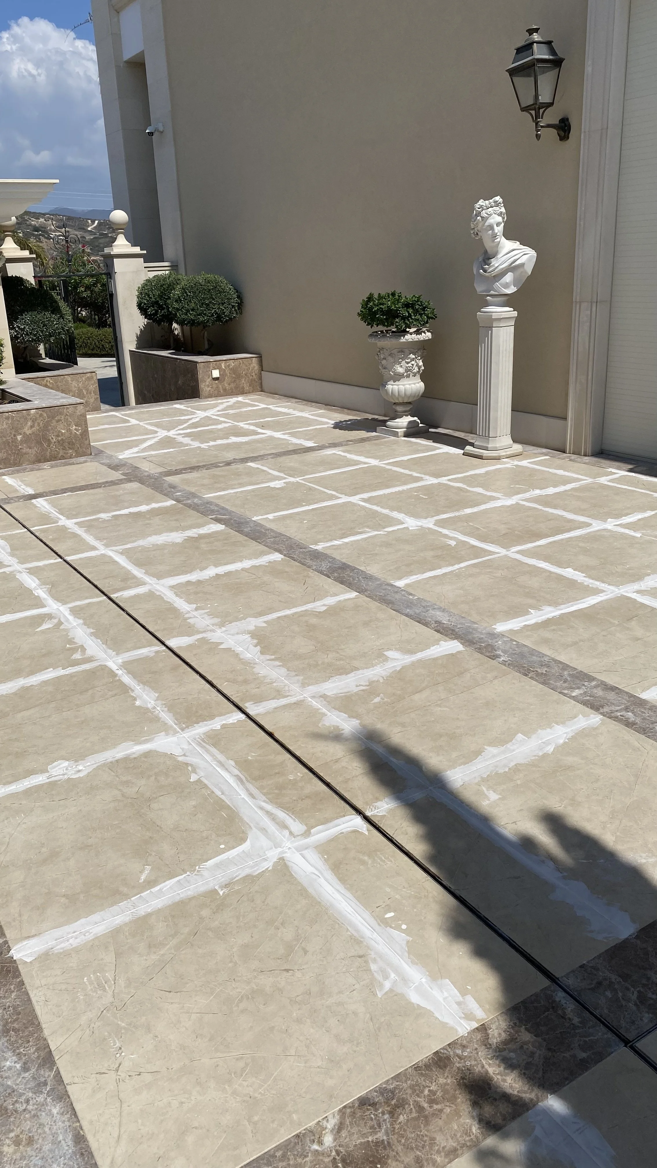 Outdoor patio with beige tiles, some are taped with painter's tape in a grid pattern, alongside decorative statues, potted plants, and a wall-mounted lantern.
