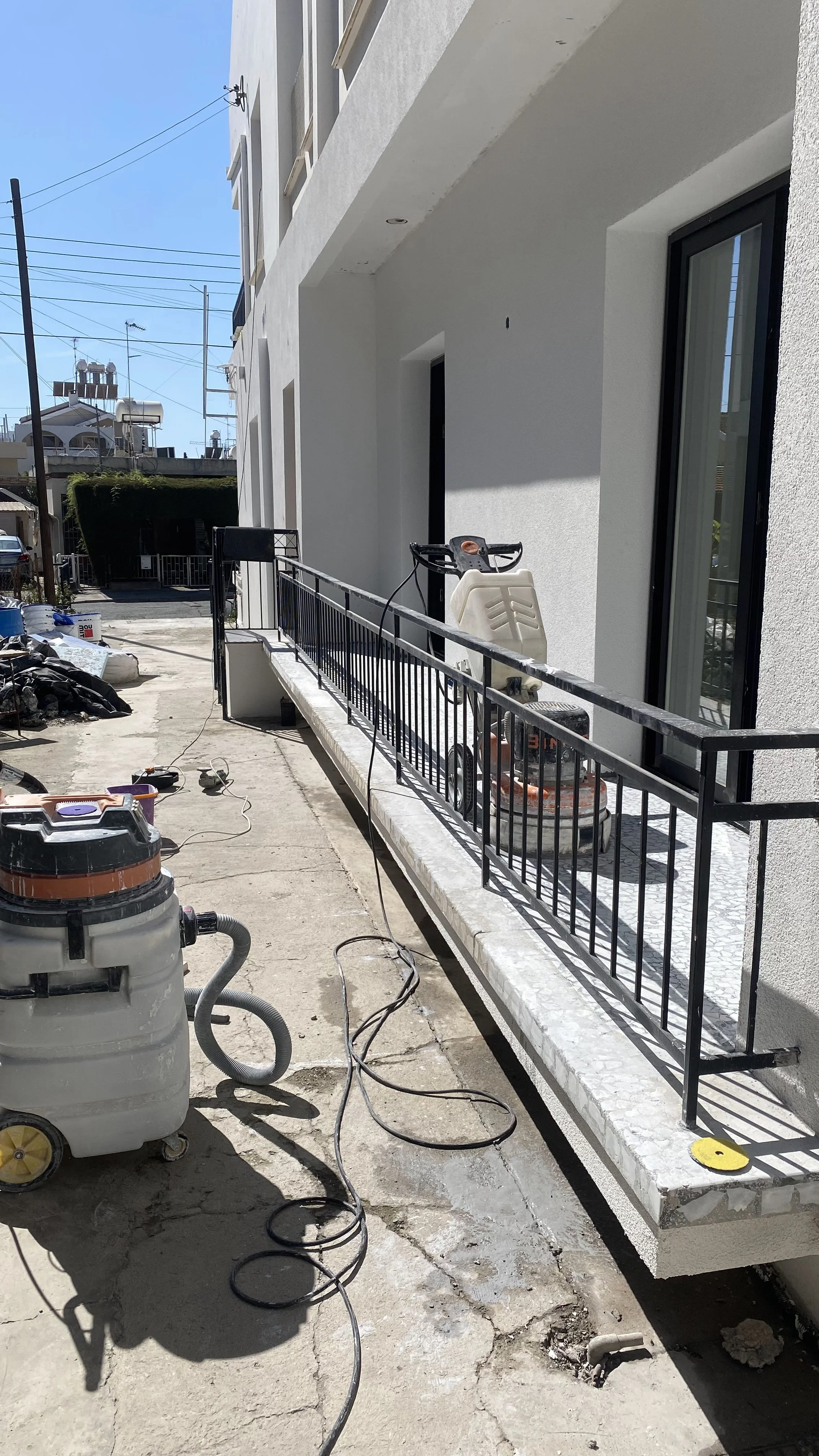 Building exterior with white walls, black balcony railing, window, and construction tools including wet vacuum, sander, and cords. Construction work ongoing.