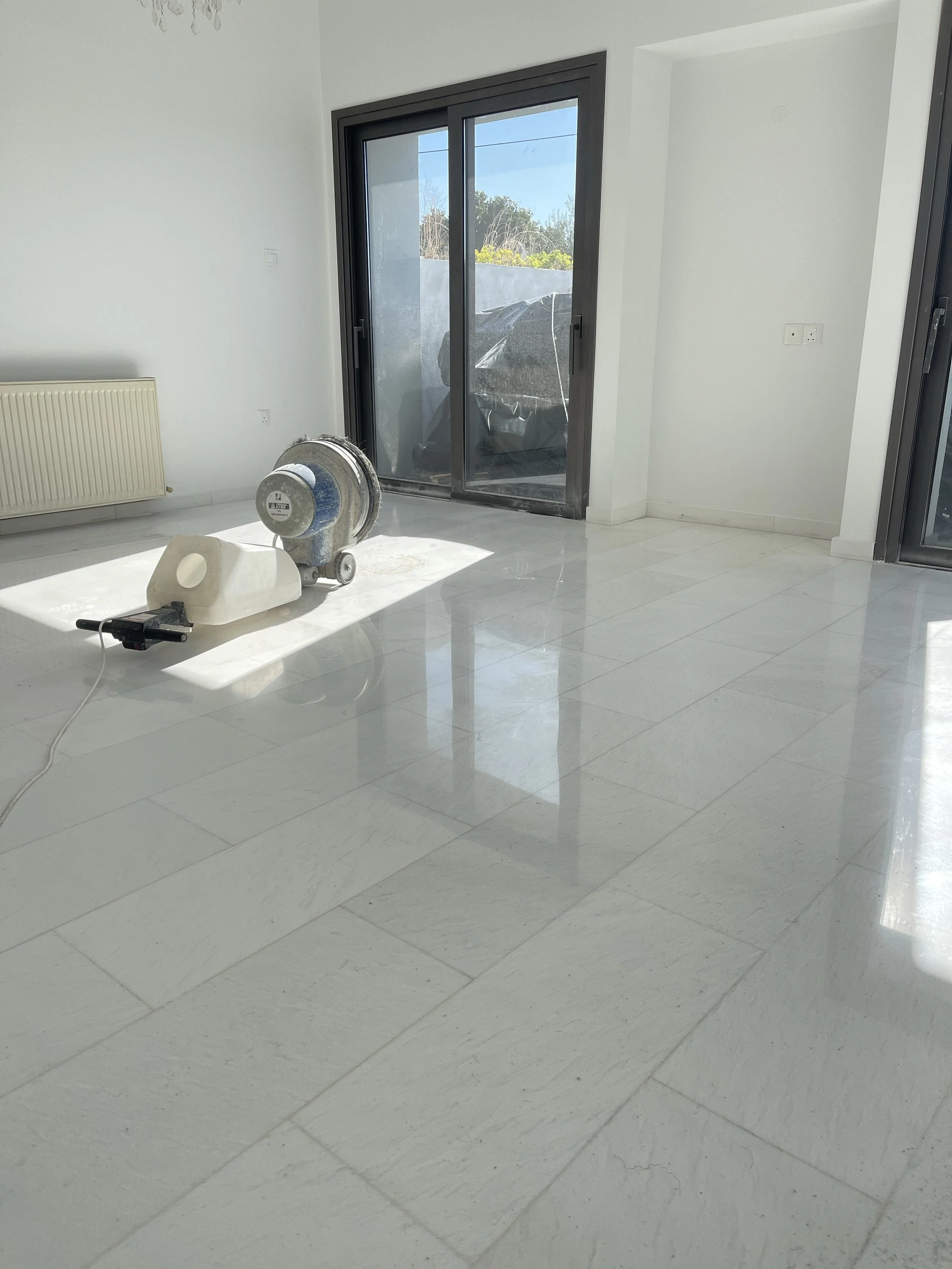 Empty room with white tiled floor, sliding glass door leading outside, radiator on the left wall, and construction equipment on the floor.