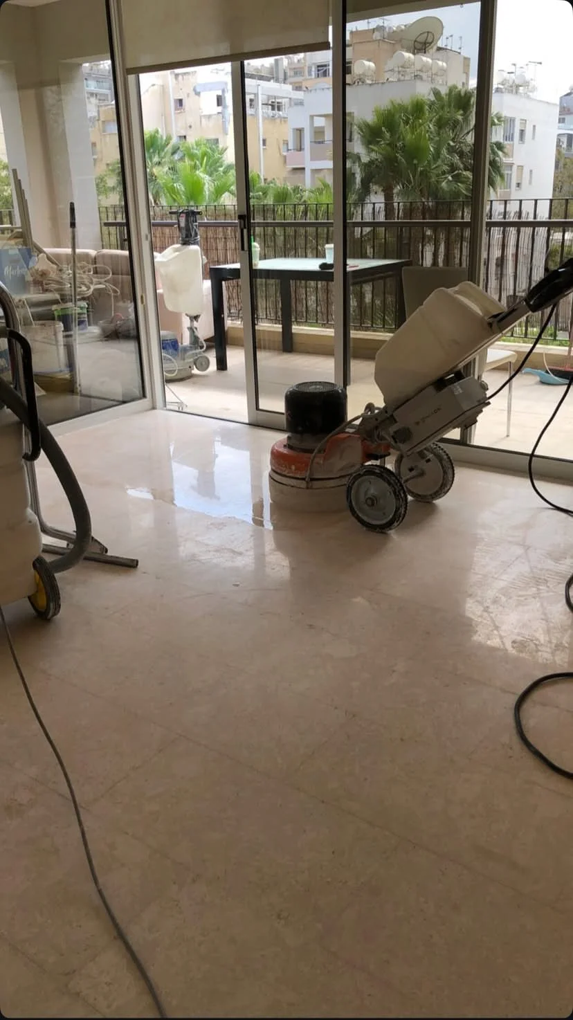 A room with a marble floor being cleaned, with a floor buffer and vacuum cleaner, and a balcony with trees and apartment buildings outside.