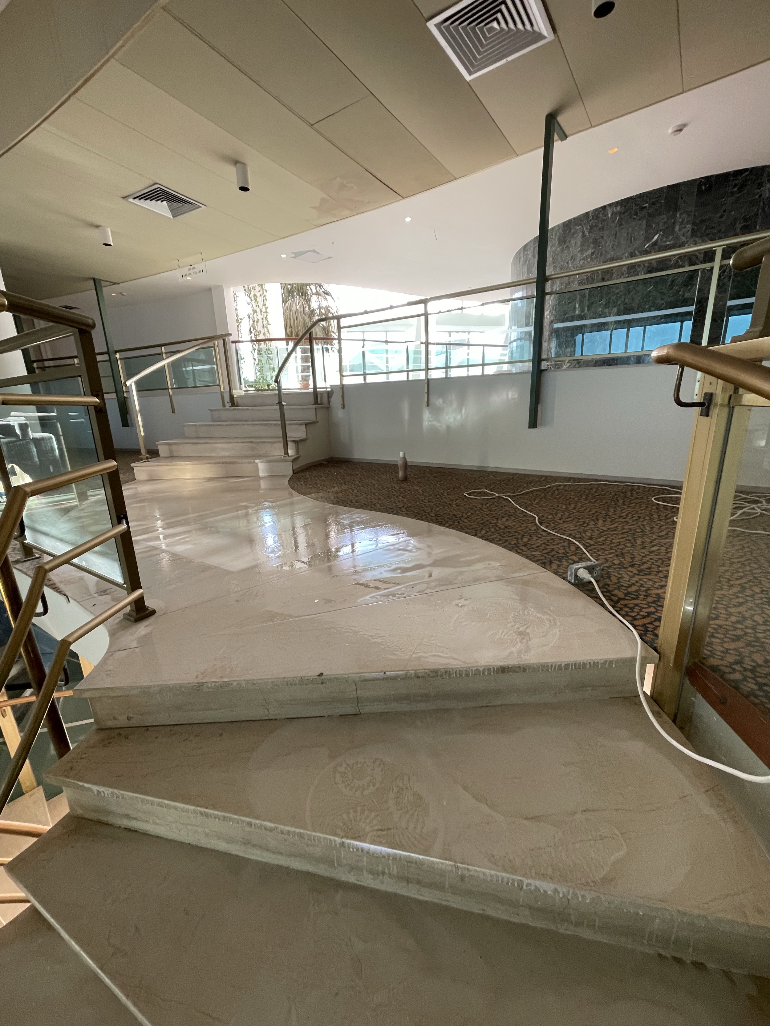 Interior view of a staircase area with marble stairs, glass railings, a curved wall with dark marble finish, and large windows allowing natural light. There are some electrical cords and a bottle on the carpeted area.