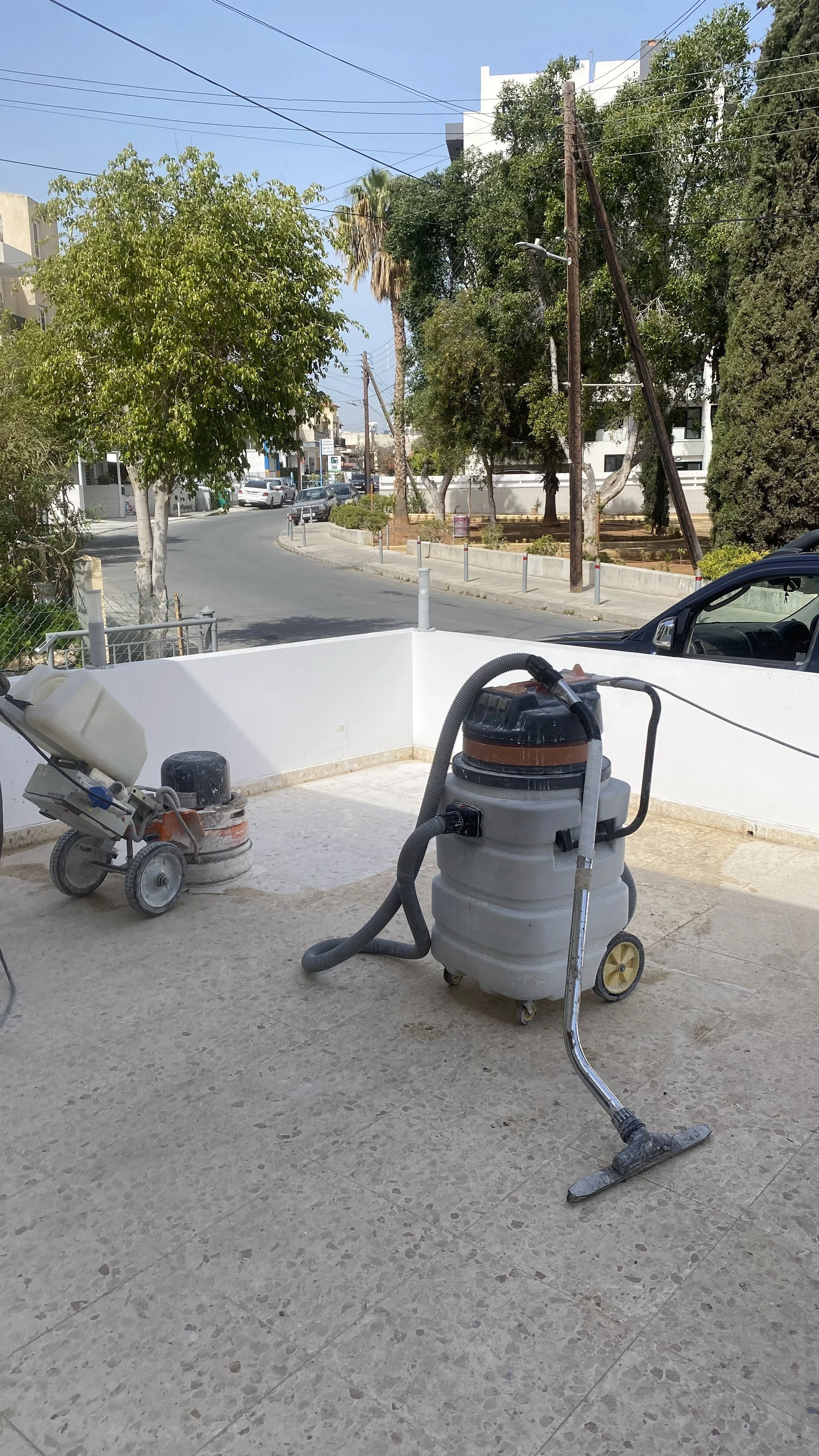 Street view with trees, parked cars, utility poles, and a white building. Construction equipment on a tiled terrace, including a vacuum and a concrete grinder.