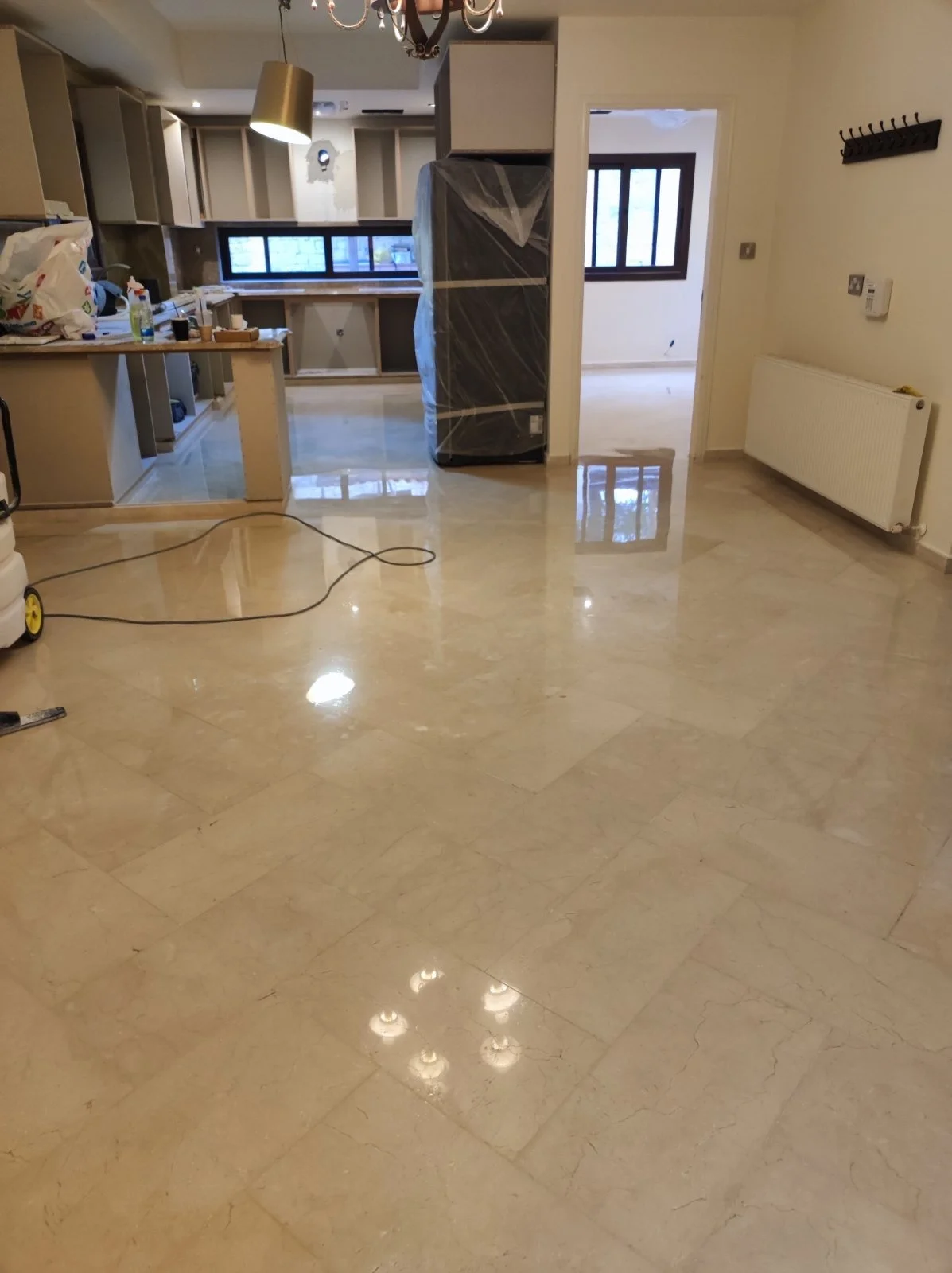 Empty room with beige marble floor, partially furnished kitchen area, window, radiator, and a doorway, with construction or renovation materials and tools present.