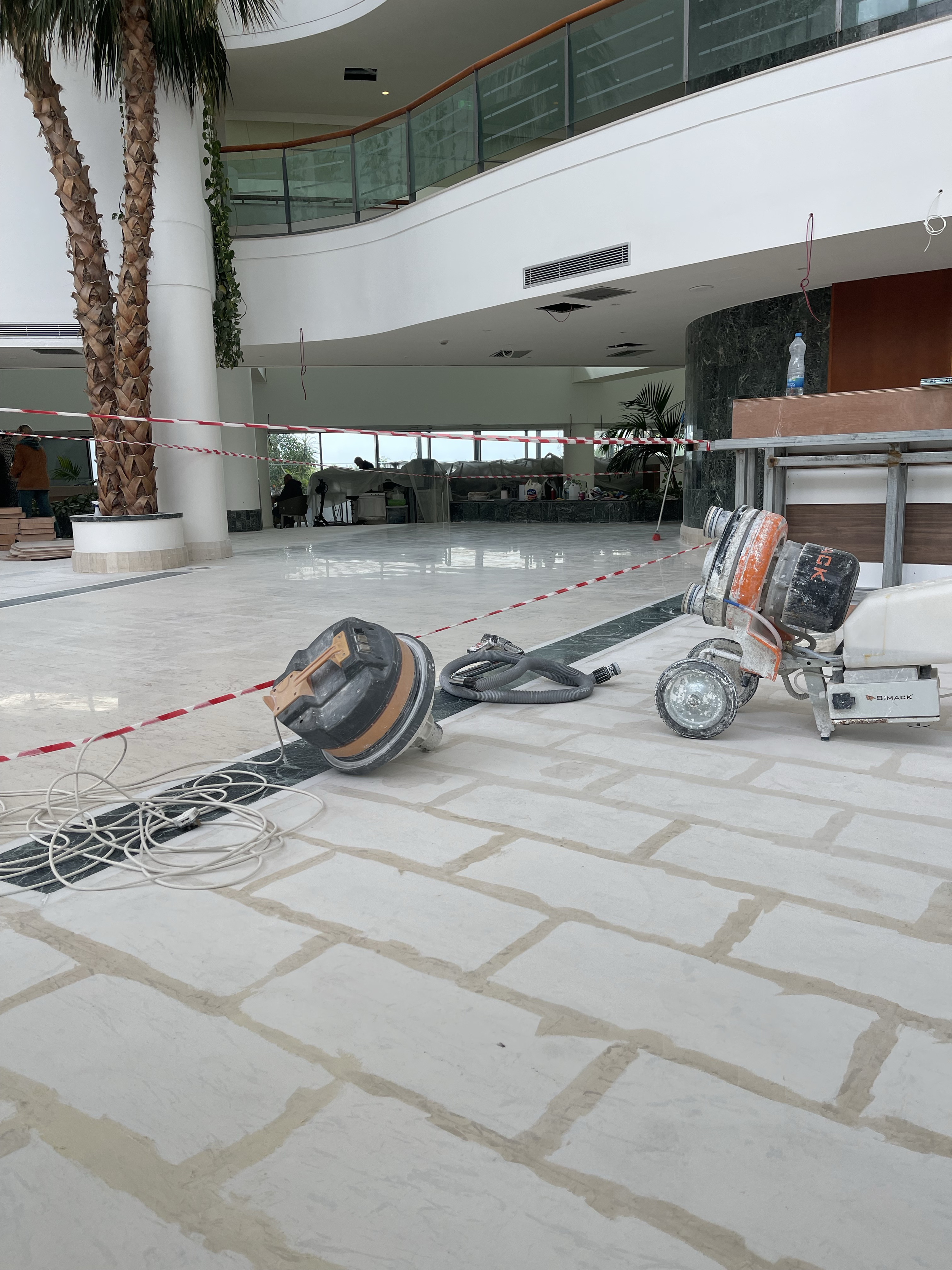 Interior of a building under construction or renovation with construction equipment, tools, and caution tape. A palm tree and large windows are visible in the background.