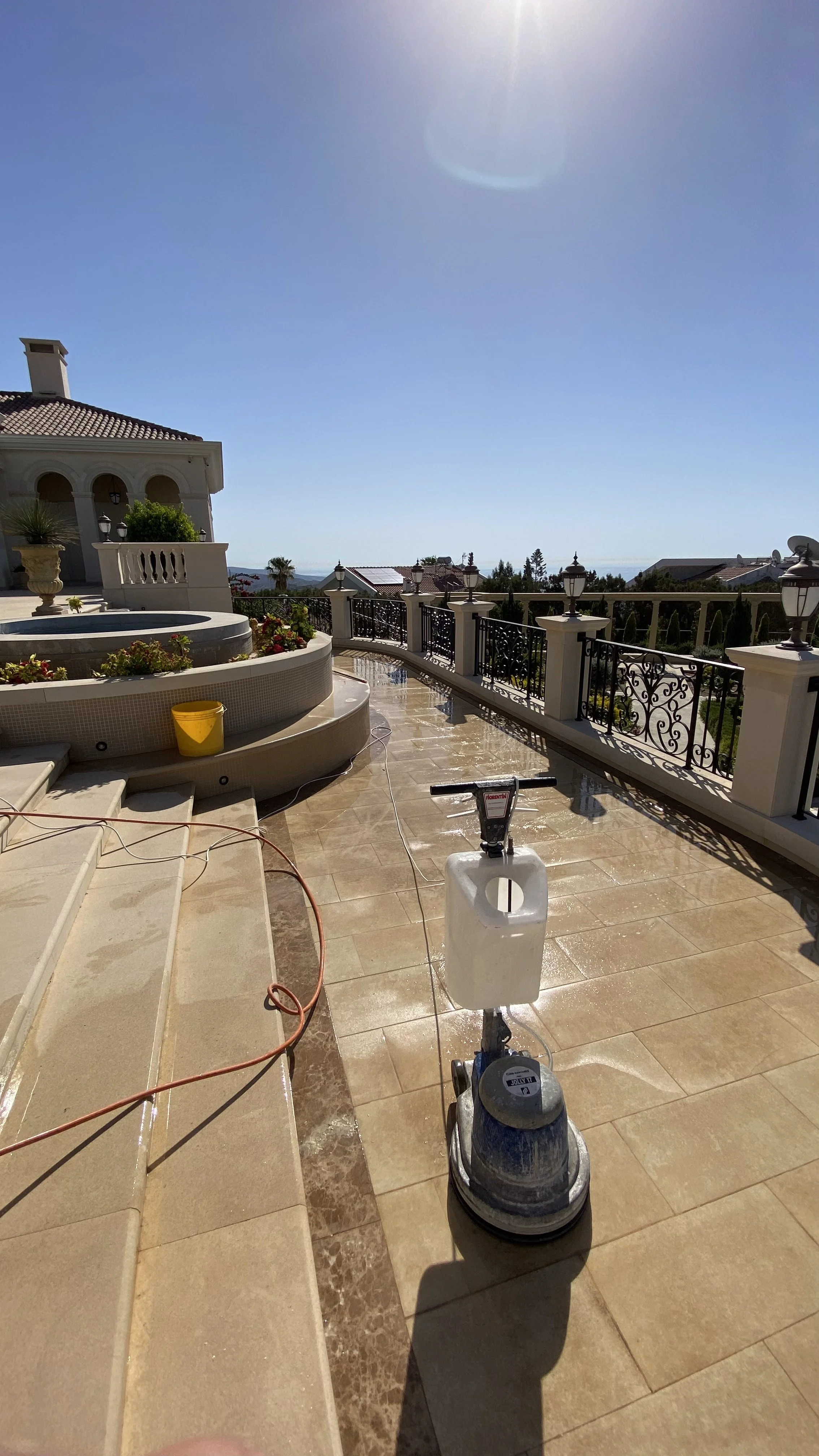 A luxurious backyard patio under construction, with a stone hot tub, potted plants, a yellow bucket, a power tool, and a floor polishing machine. The patio is fenced, overlooking houses and trees in the distance with a clear sunny sky.