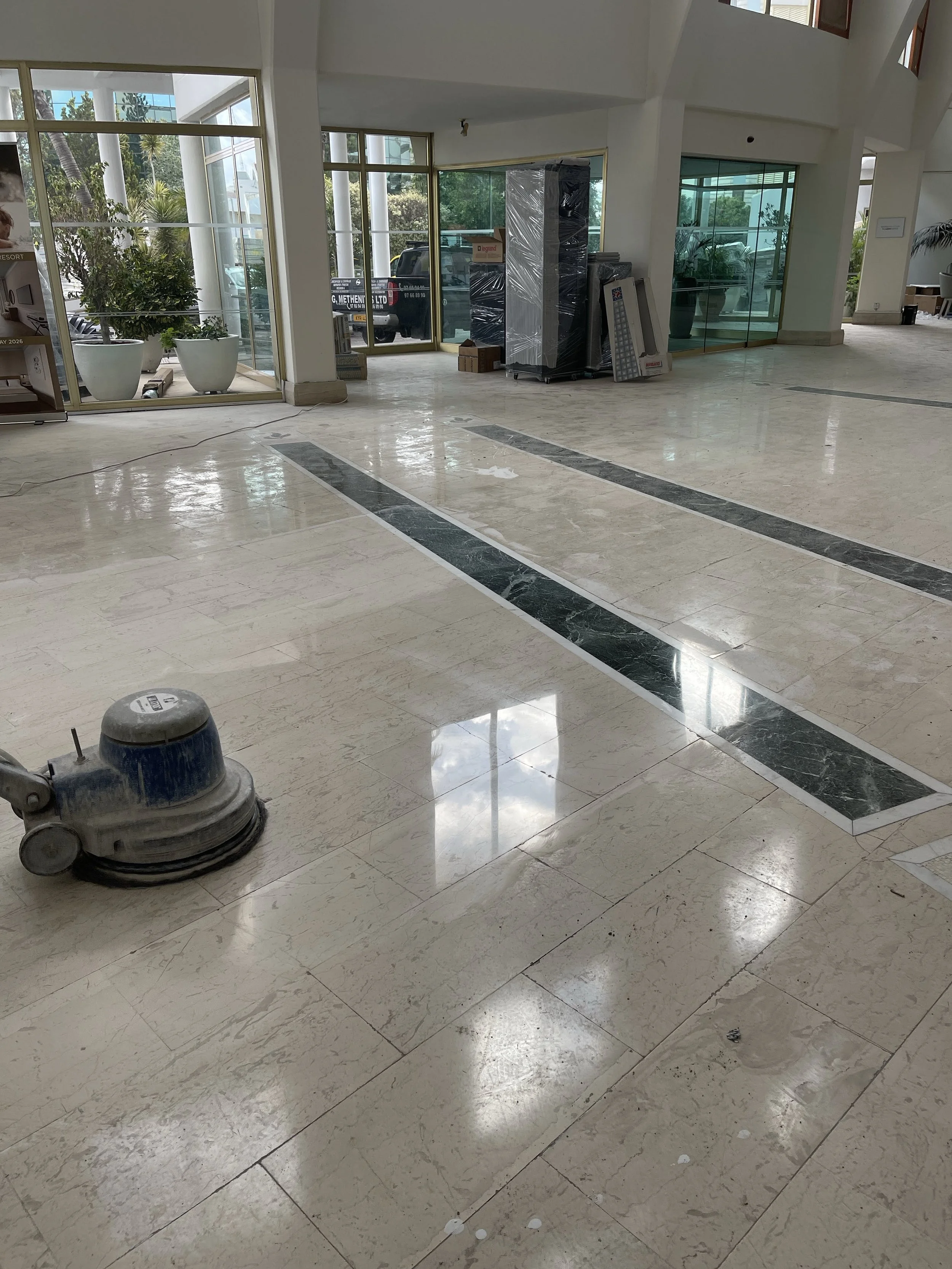 Interior of a building under construction or renovation, with a floor cleaning machine, large potted plants outside the glass entrance, and construction materials and packaging stacked near the glass doors.