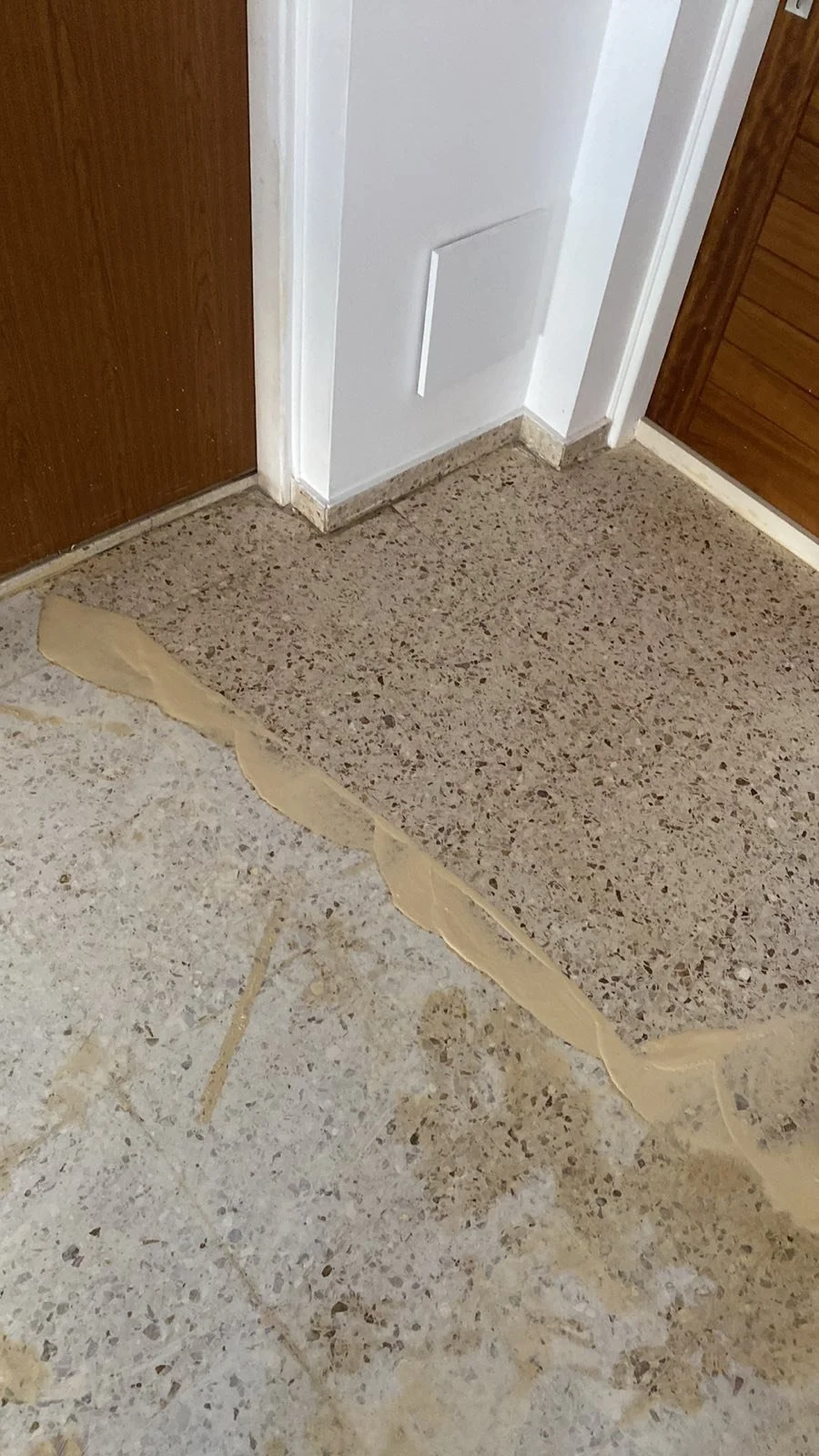 Floor with beige tiles and a section of brown speckled flooring, with a patch of beige construction or repair material at the edge of the brown section near a white wall and wooden doors.