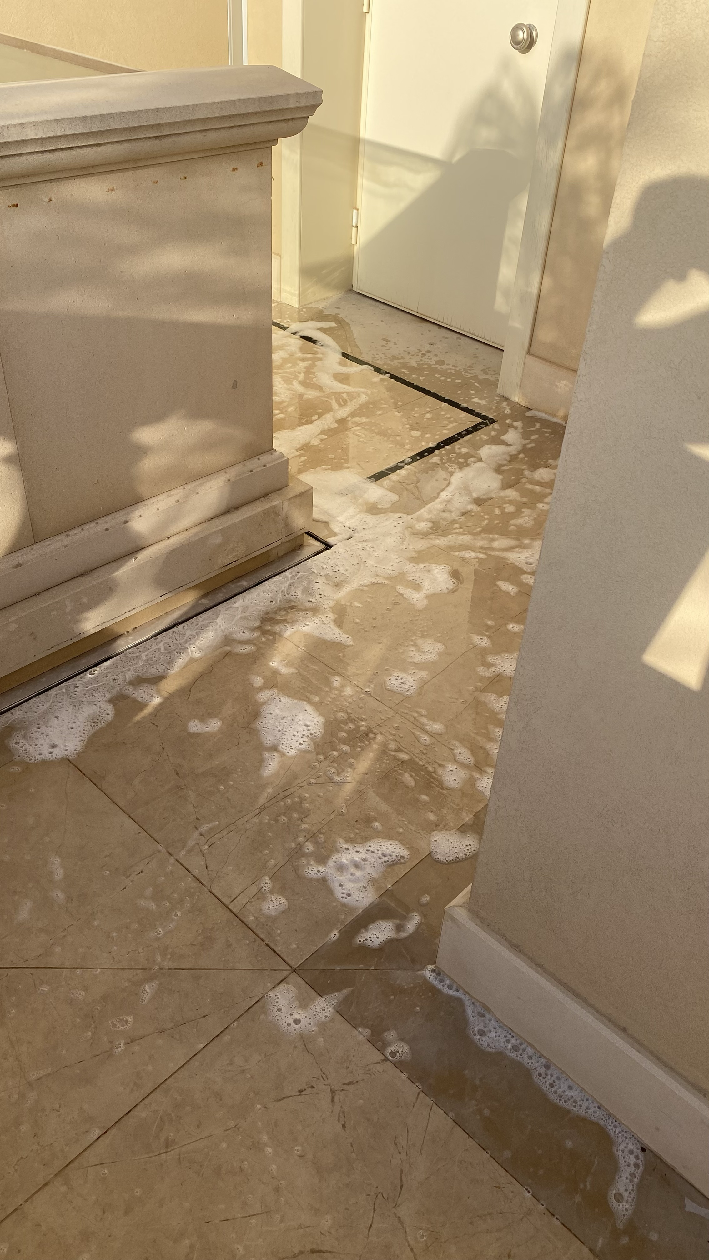 Foamy water on a beige tiled bathroom floor, flowing out from under a partially open door.