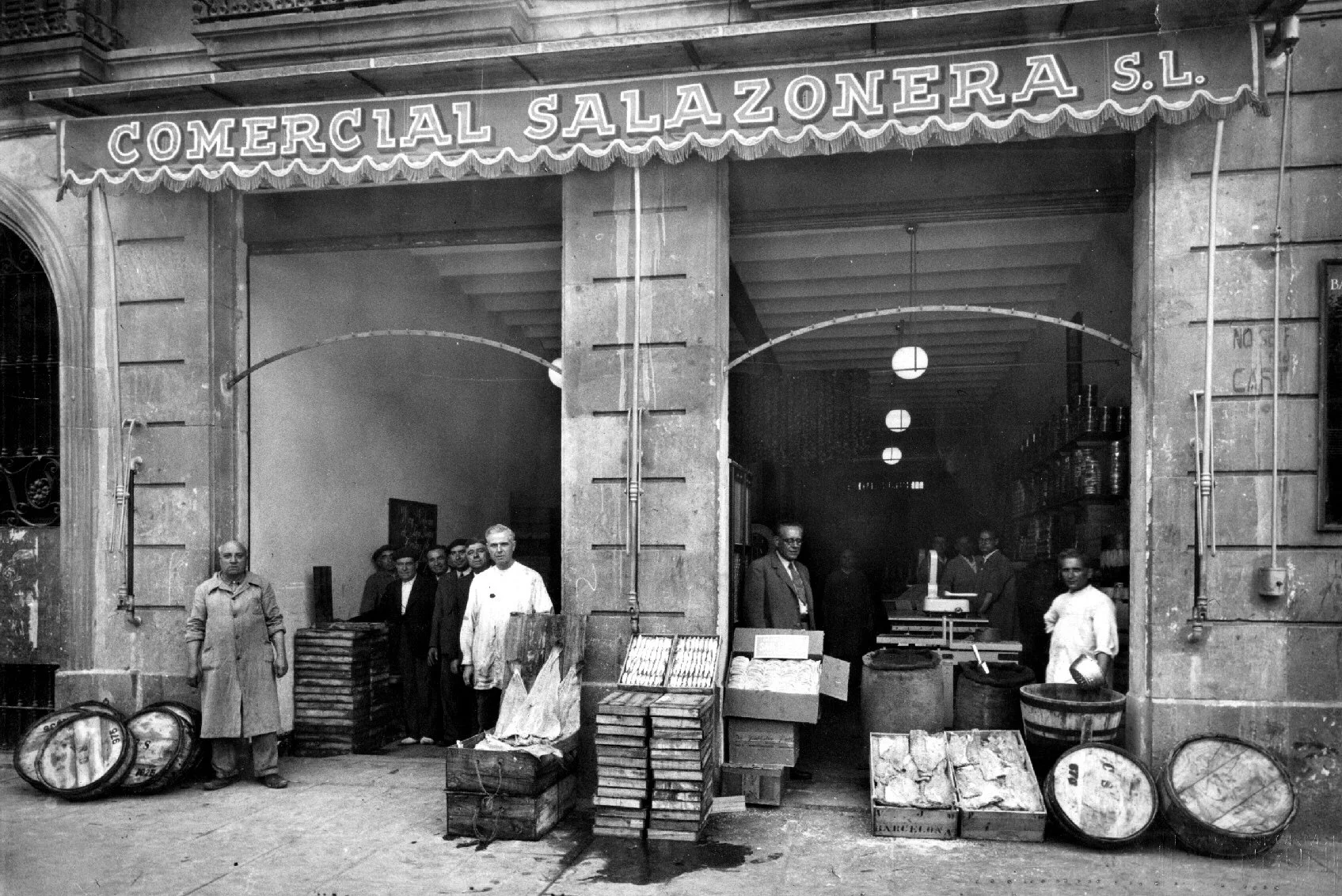 Black and white photo of a storefront with a sign reading 'COMERCIAL SALAZONERA S.L.' above the entrance. Several people stand outside, some behind a table with barrels and wooden crates, and others in the background inside the shop.